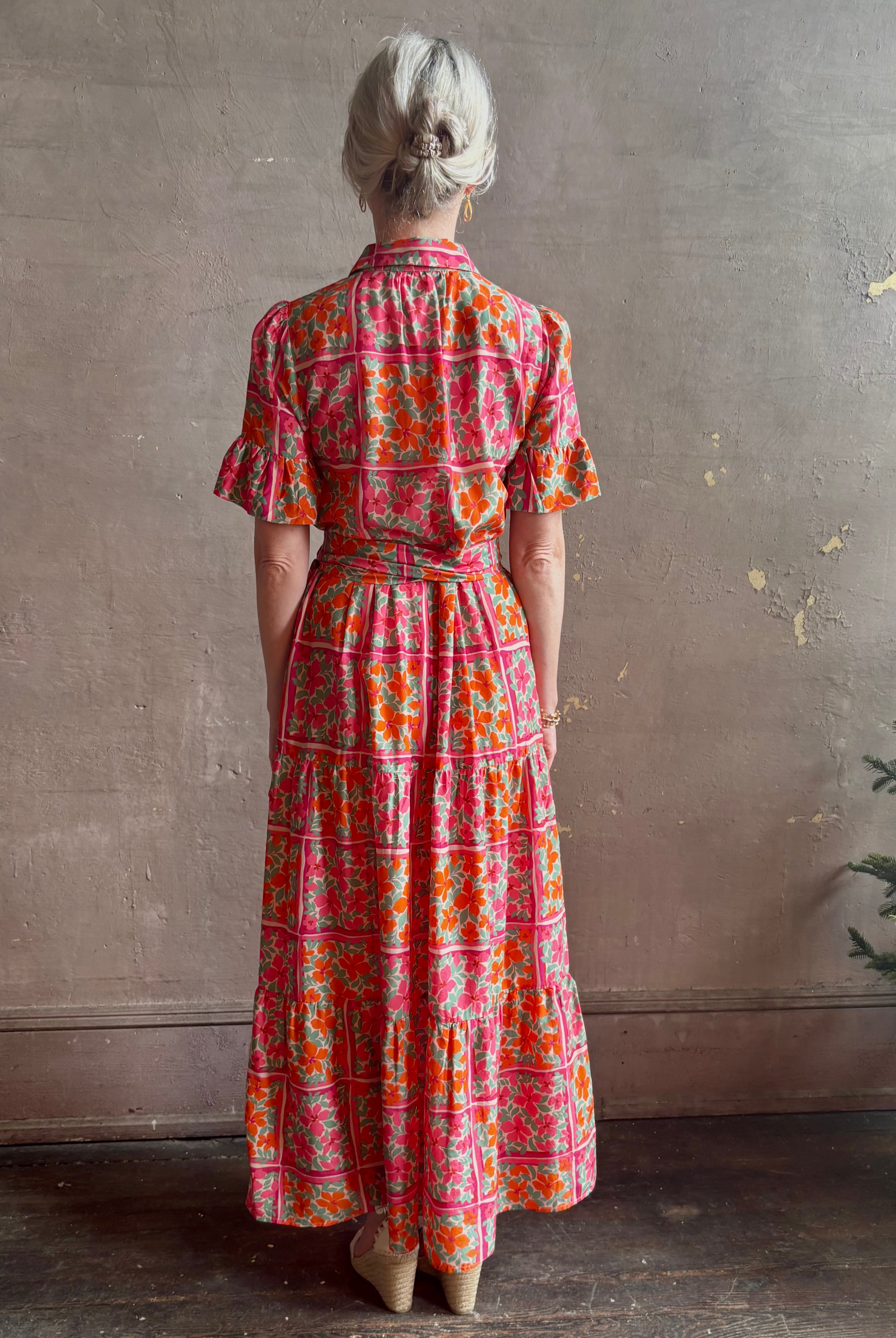 Mature woman wearing Martine maxi Dress from Trovata rendered in the Botanic Tile pink and orange floral print and crafted from a luxurious blend of cotton and silk with button front, collar, short sleeves and matching optional self tile belt