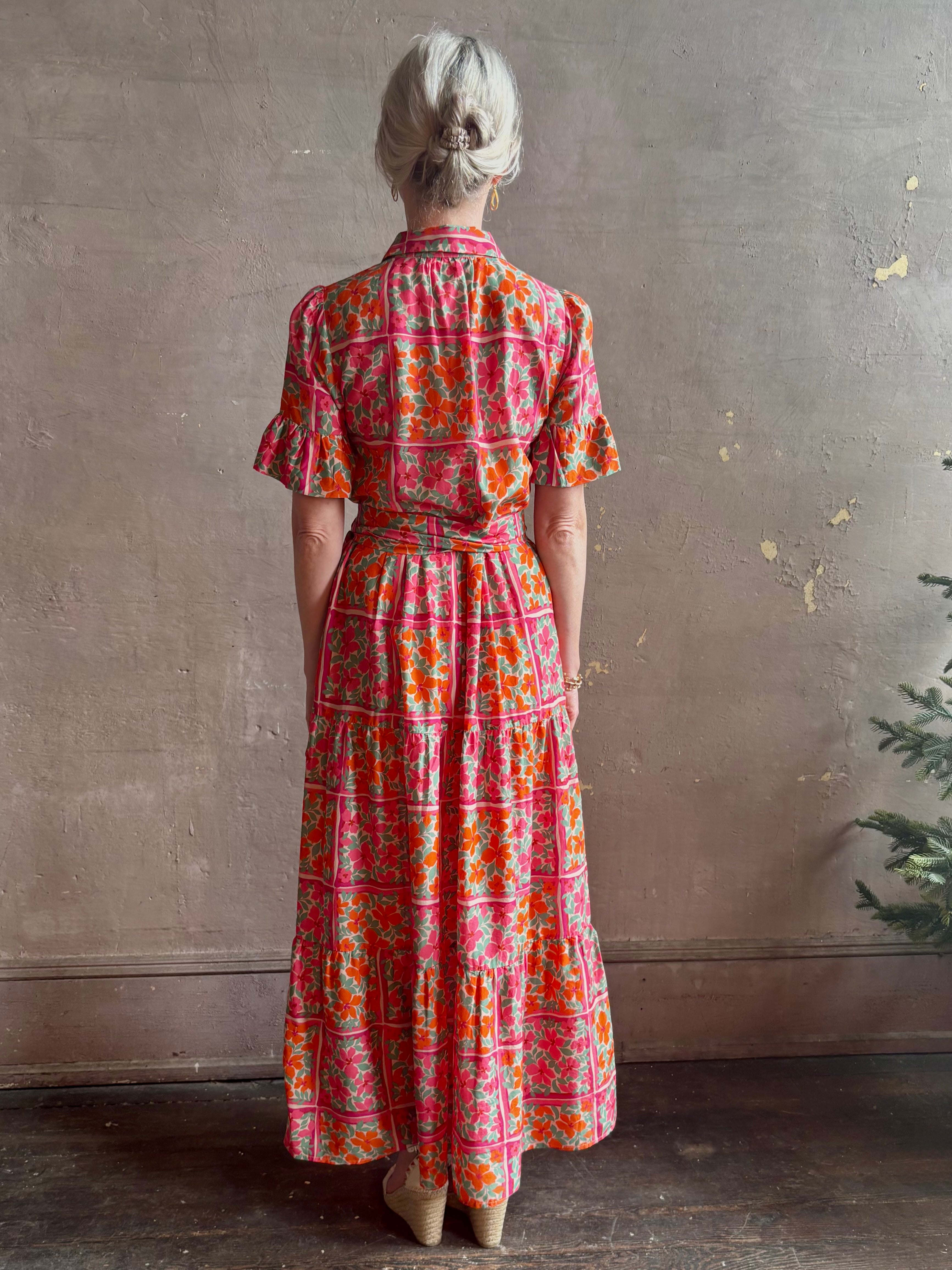 Mature woman wearing Martine maxi Dress from Trovata rendered in the Botanic Tile pink and orange floral print and crafted from a luxurious blend of cotton and silk with button front, collar, short sleeves and matching optional self tile belt