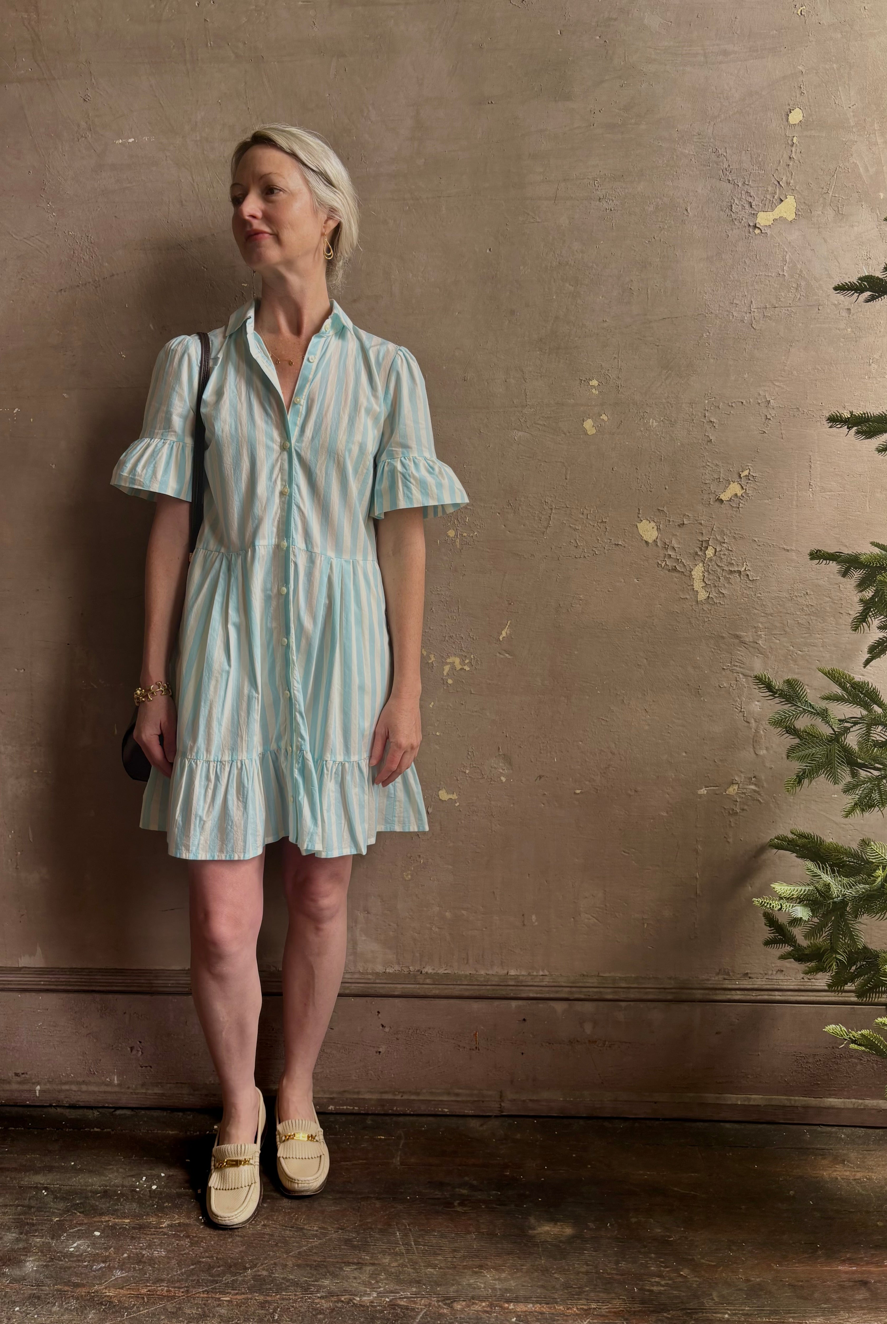 Mature woman wearing Martinique Dress from Trovata rendered in a cream and aqua Stripe and crafted from cotton poplin with ruffle trimmed short sleeves, collared button front short dress and co-ordinating fabric tie belt