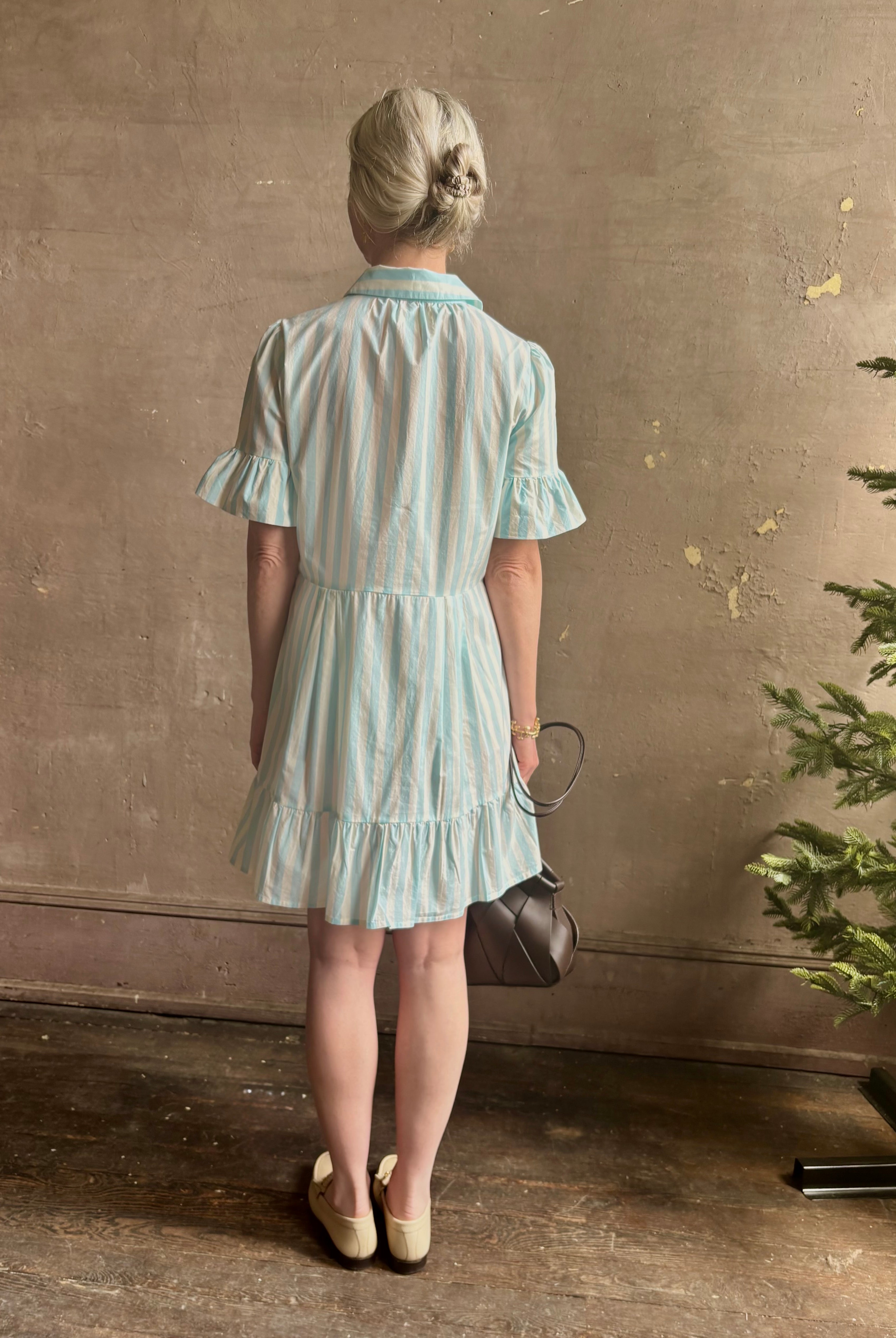 Mature woman wearing Martinique Dress from Trovata rendered in a cream and aqua Stripe and crafted from cotton poplin with ruffle trimmed short sleeves, collared button front short dress and co-ordinating fabric tie belt