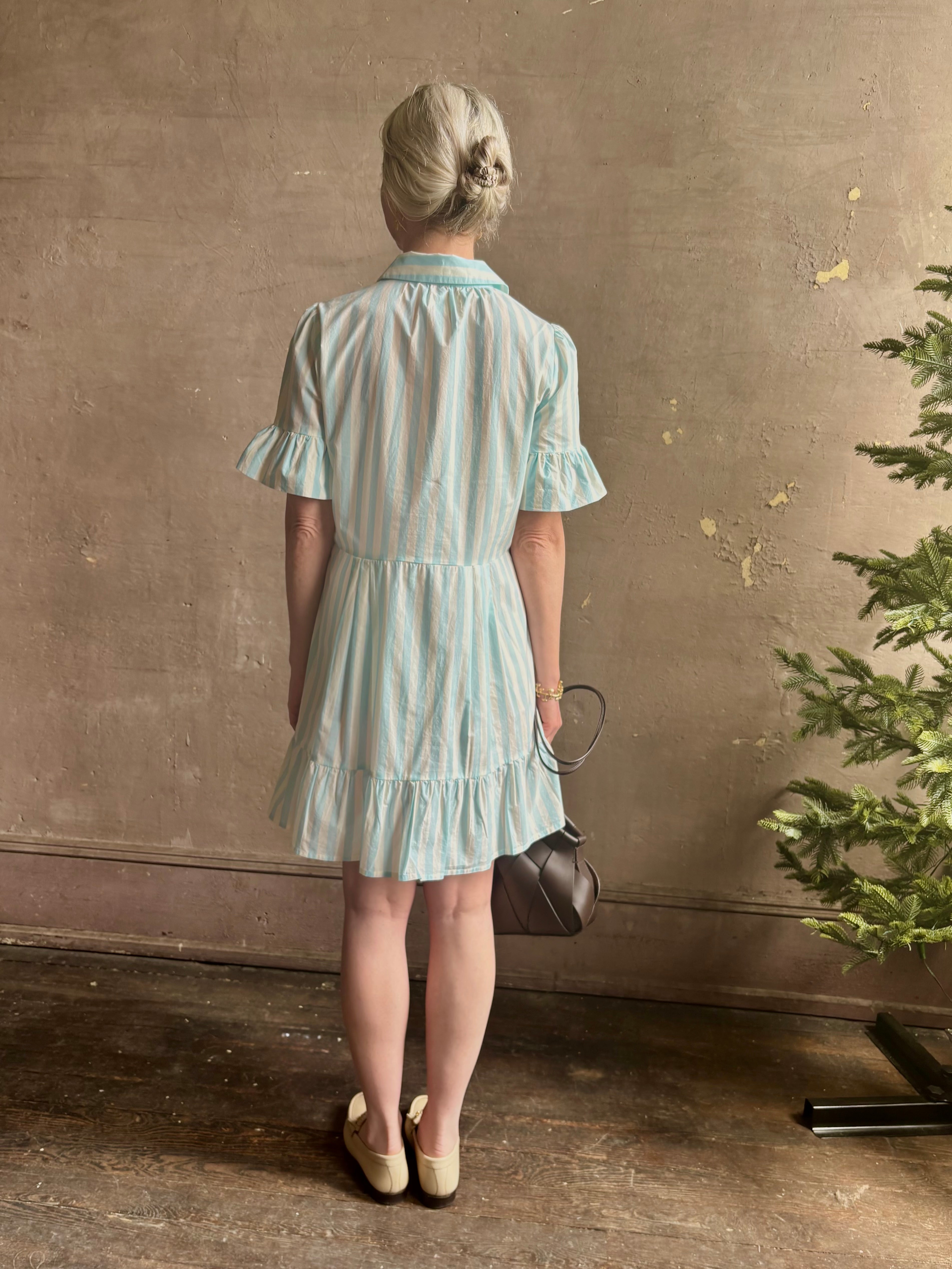Mature woman wearing Martinique Dress from Trovata rendered in a cream and aqua Stripe and crafted from cotton poplin with ruffle trimmed short sleeves, collared button front short dress and co-ordinating fabric tie belt