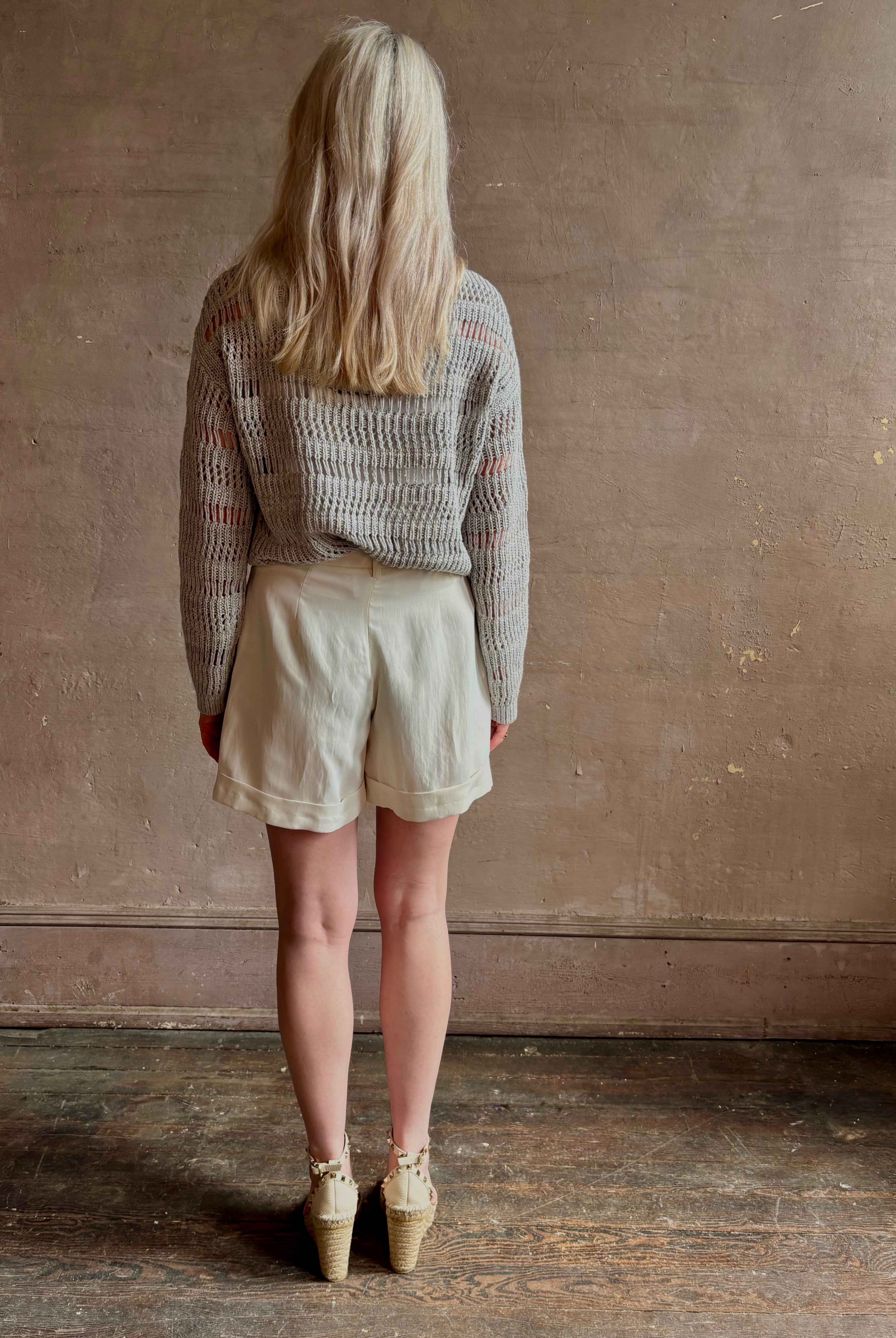 Mature woman wearing elevated tailored Cisco Shorts from TWP rendered in a soft bone white and crafted in the USA using a viscose and linen blend fabric with side seam pockets, belt loops, single front pleat and cuffed at the bottom