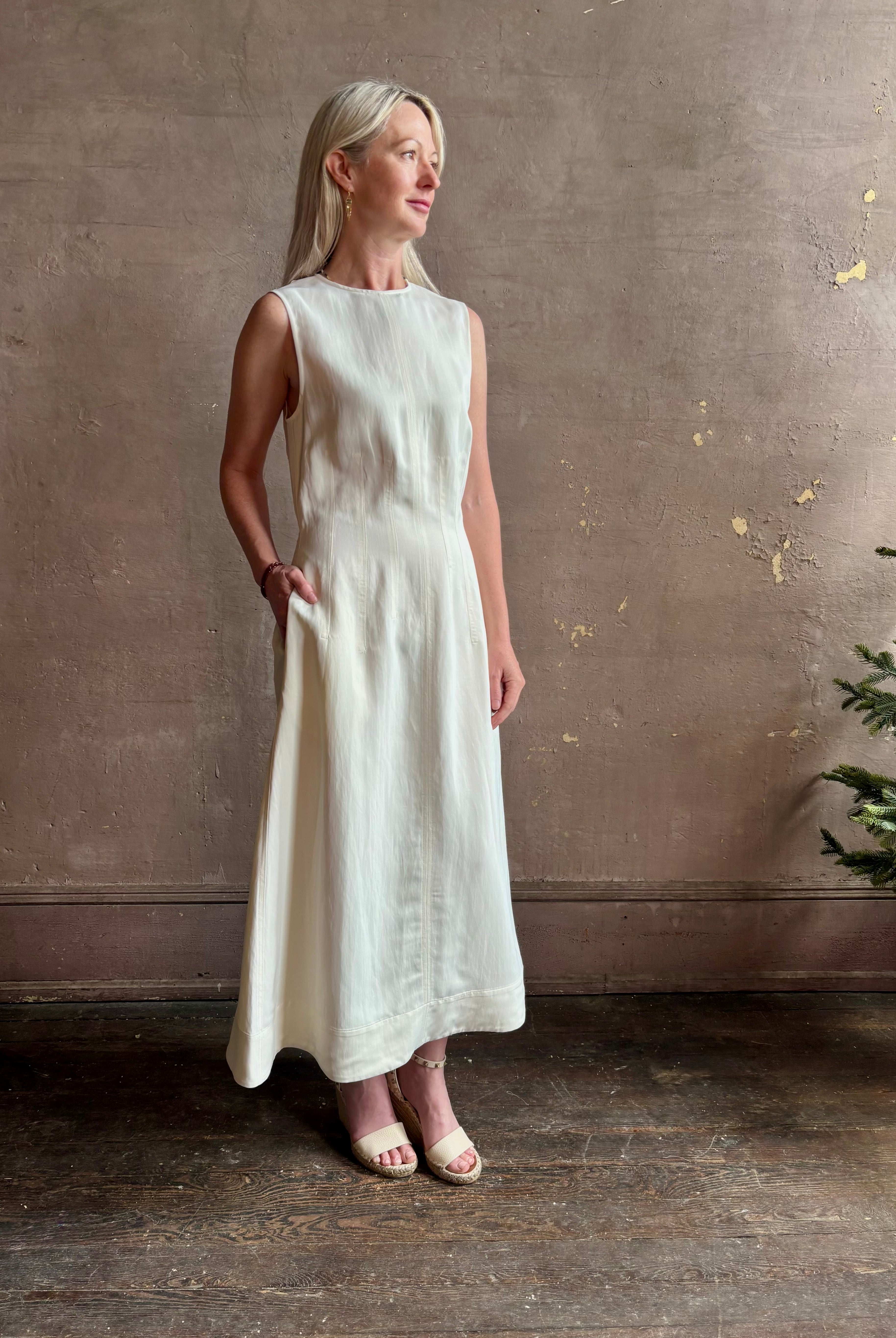 Mature woman wearing elegant Crew Neck maxi A-Line Dress with Darts from TWP rendered in a soft off white and crafted in the USA from a viscose and linen fabric blend with side seam pockets and sleeveless