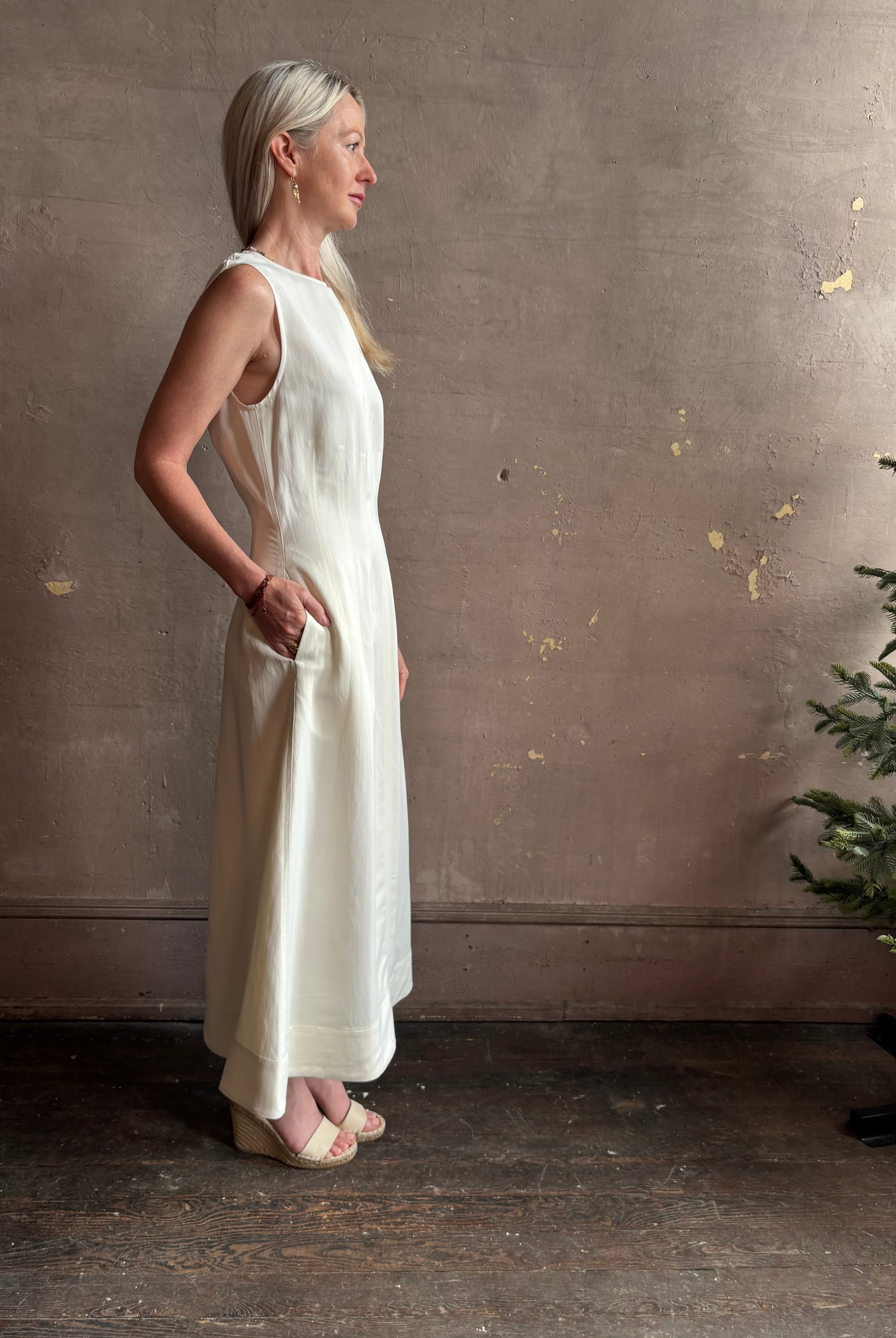 Mature woman wearing elegant Crew Neck maxi A-Line Dress with Darts from TWP rendered in a soft off white and crafted in the USA from a viscose and linen fabric blend with side seam pockets and sleeveless
