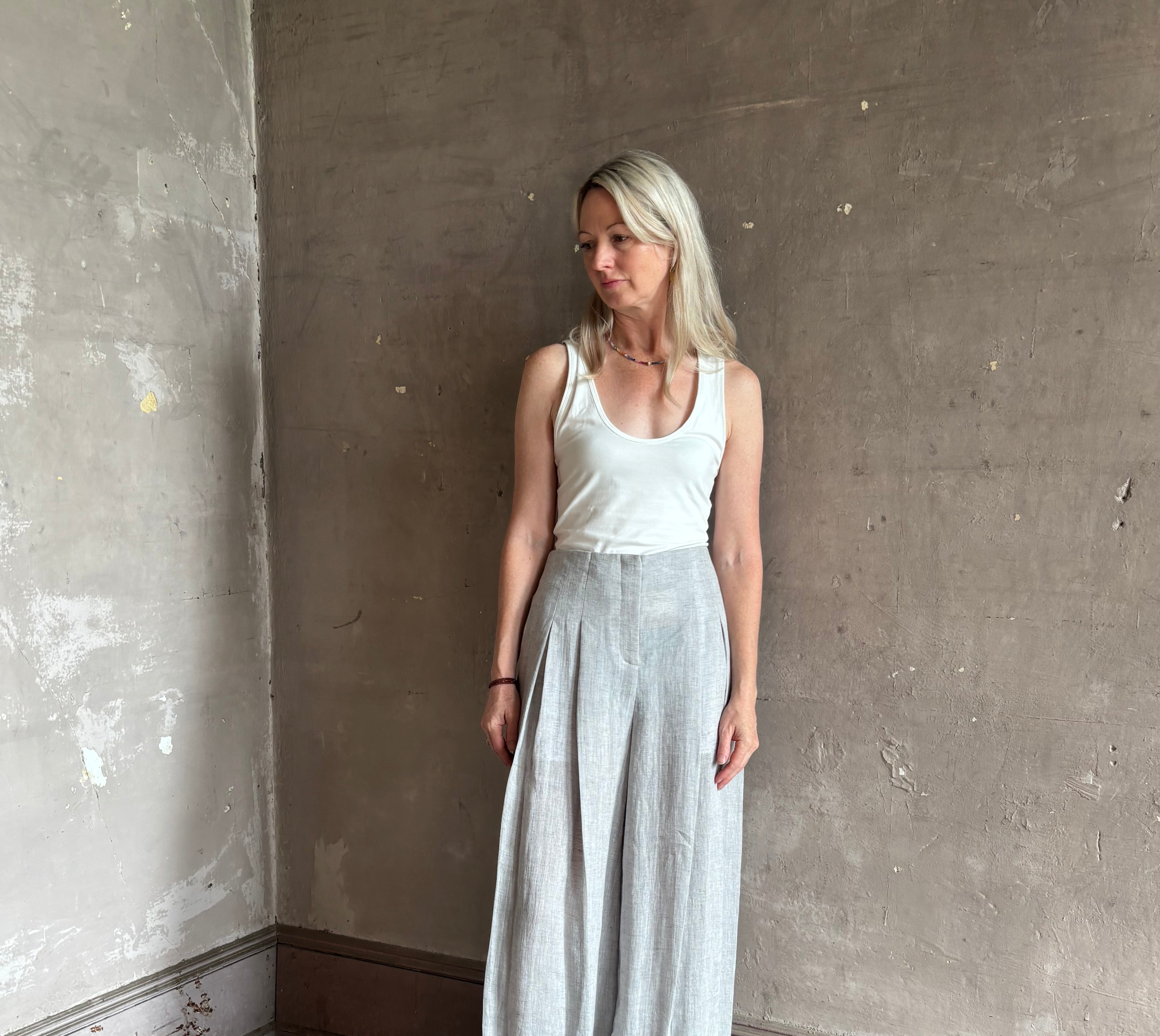 Mature woman wearing effortless women's designer Drew wide leg Trouser from TWP rendered in a versatile light heather grey and crafted from light weight hemp with high-rise, drop waist pleats, and side seam pockets, and made in the USA