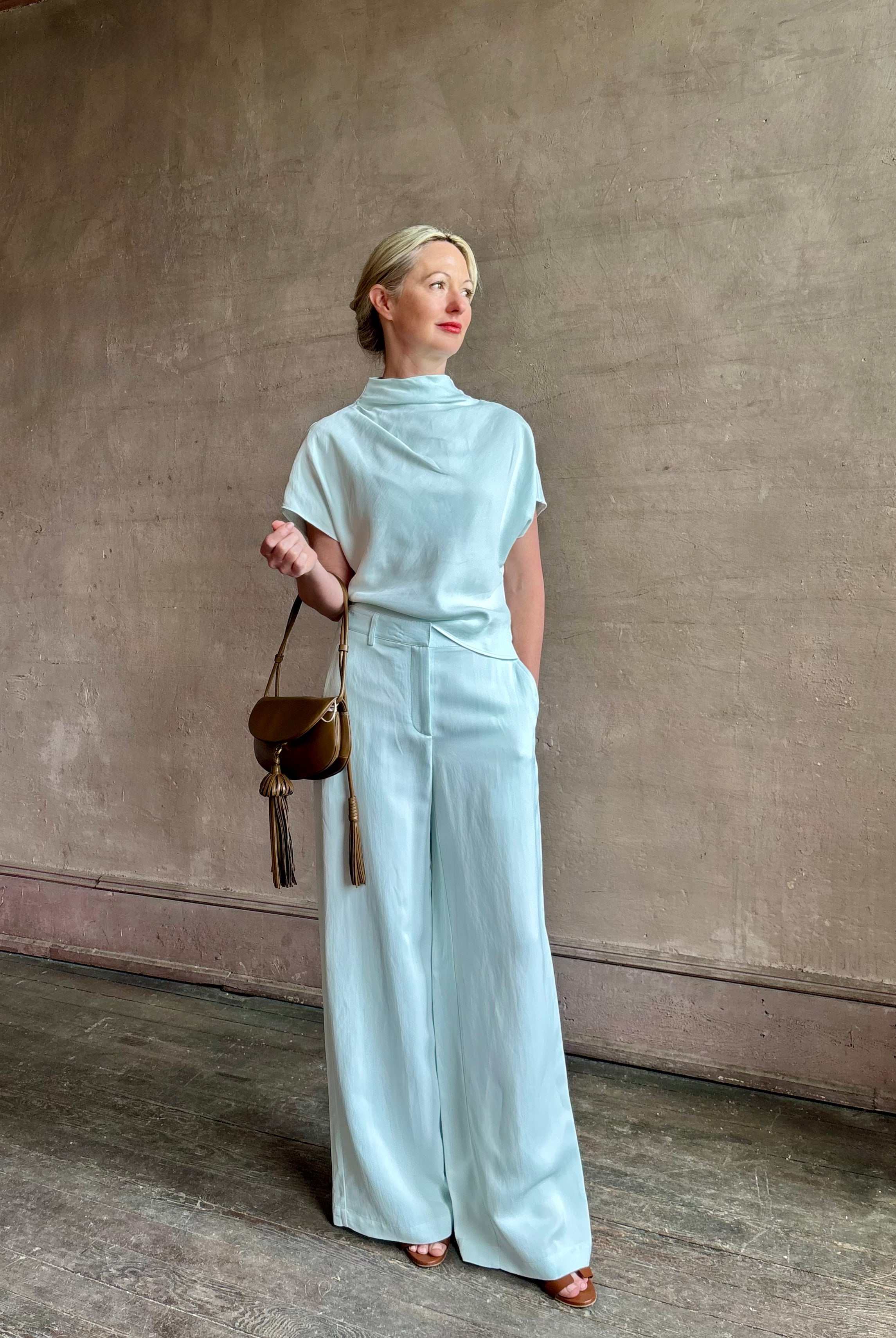 Mature woman wearing the Dylan Pant from TWP rendered in a sea glass blue hue and crafted from a viscose and linen blend, made in the USA, this luxury mid-rise trouser offers a wide leg silhouette with hidden seam side and back pockets