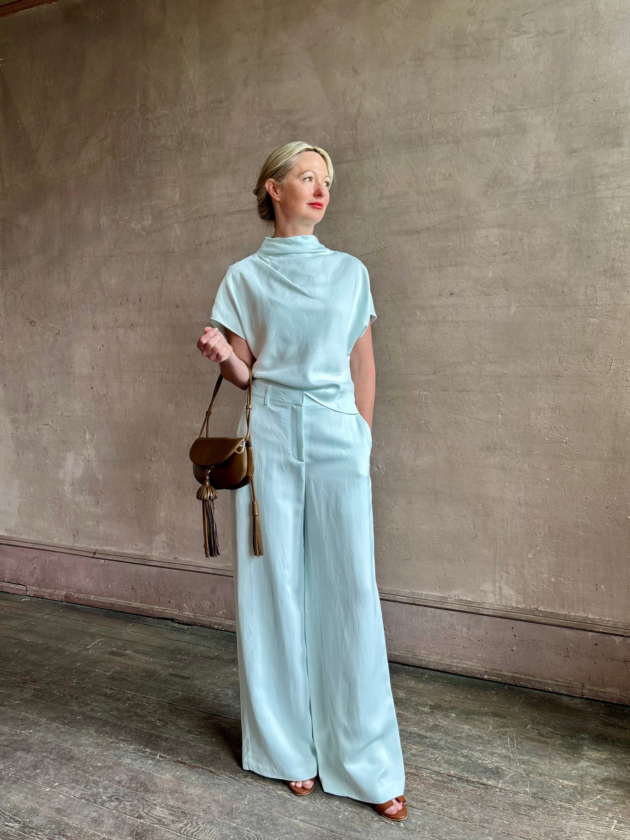 Mature woman wearing the Dylan Pant from TWP rendered in a sea glass blue hue and crafted from a viscose and linen blend, made in the USA, this luxury mid-rise trouser offers a wide leg silhouette with hidden seam side and back pockets