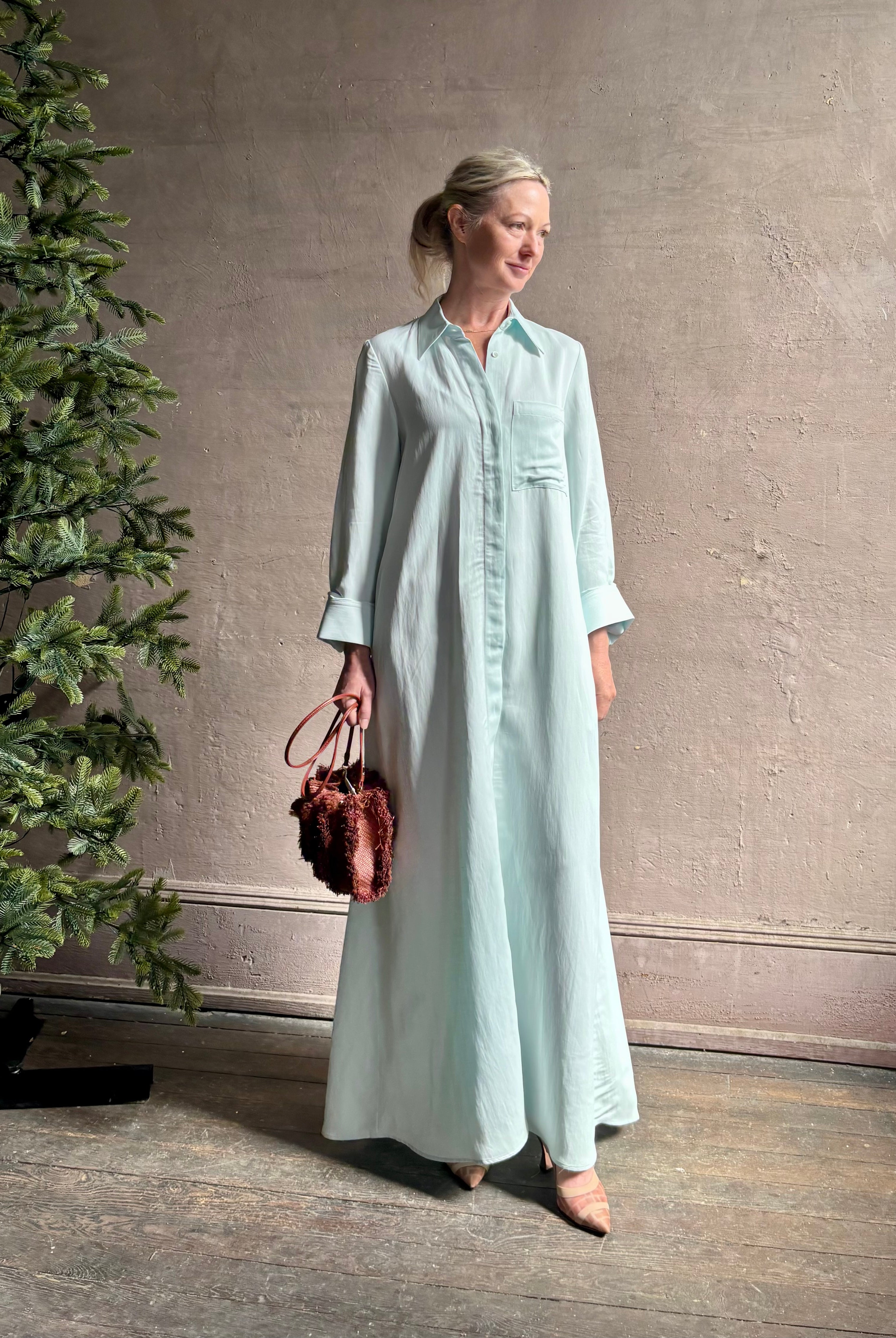 Mature woman wearing floor-length Jenny's Gown from TWP rendered in a sea glass hue and crafted from a viscose linen blend with beautiful draping, long sleeves, button front and cool girl loose fit