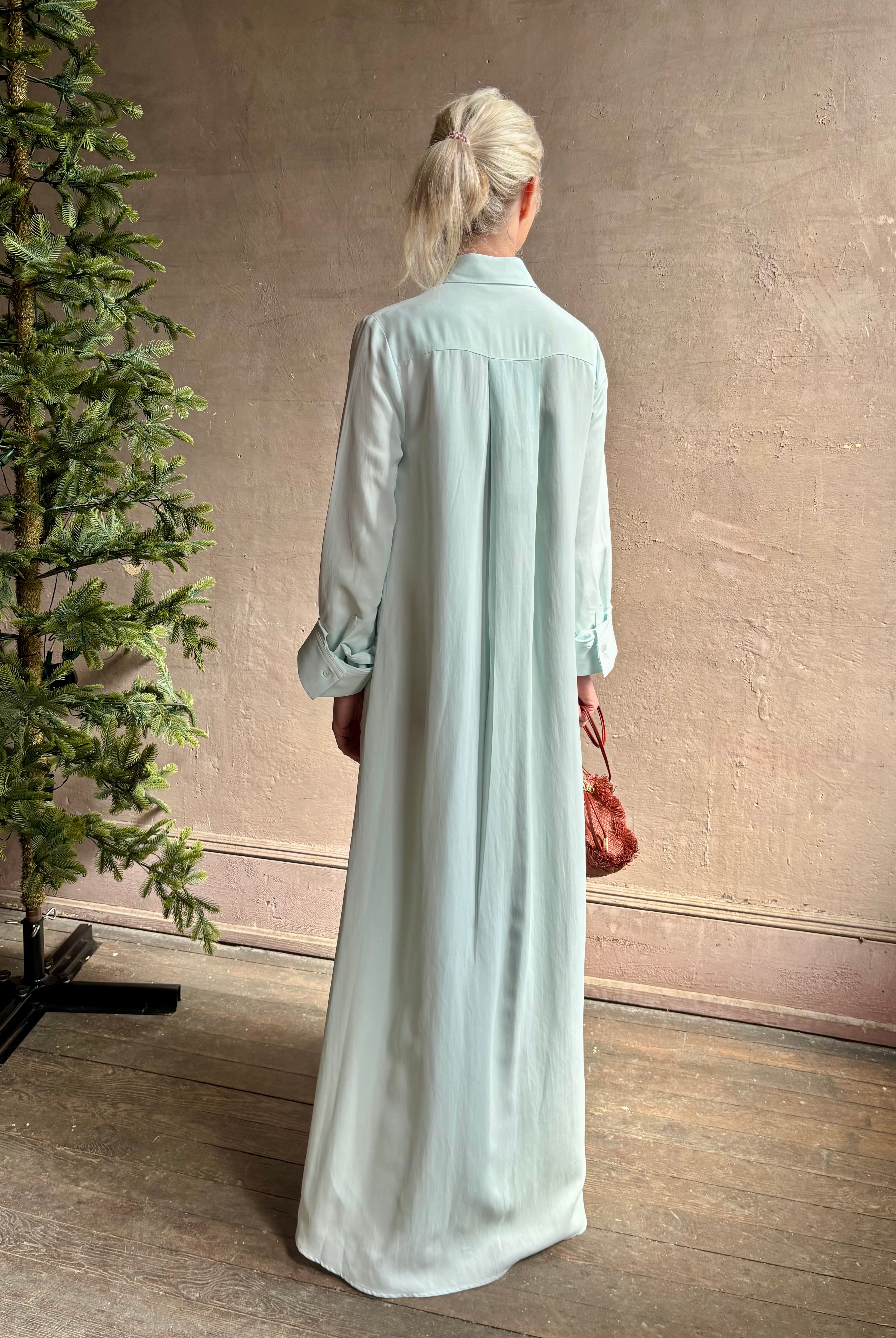 Mature woman wearing floor-length Jenny's Gown from TWP rendered in a sea glass hue and crafted from a viscose linen blend with beautiful draping, long sleeves, button front and cool girl loose fit