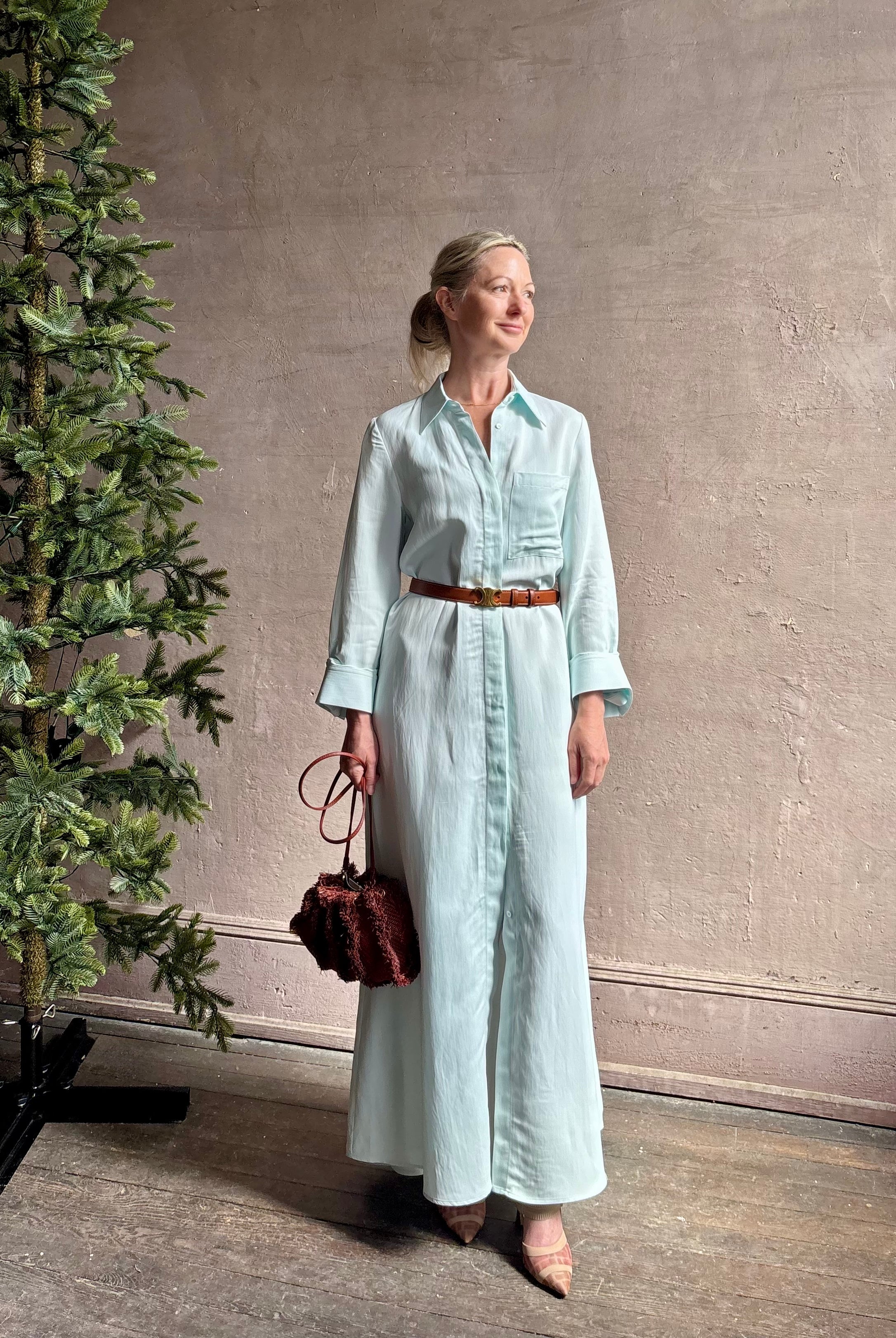 Mature woman wearing floor-length Jenny's Gown from TWP rendered in a sea glass hue and crafted from a viscose linen blend with beautiful draping, long sleeves, button front and cool girl loose fit
