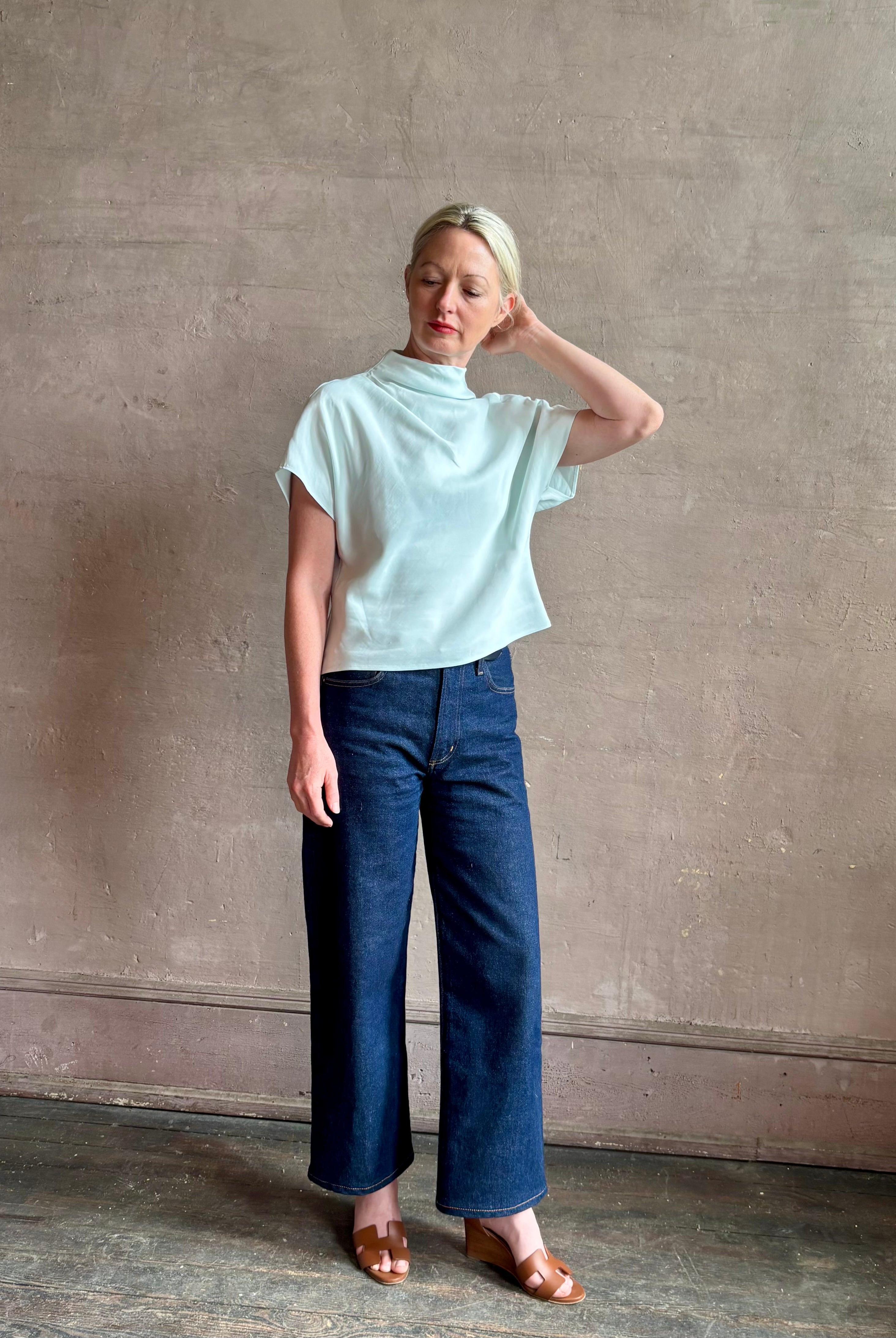 Mature woman wearing refined Mock Neck Tee Blouse from TWP rendered in a subtle sea glass blue hue and crafted from a viscose linen blend made in the USA, with short sleeves, asymmetrical design and quality fabric draping
