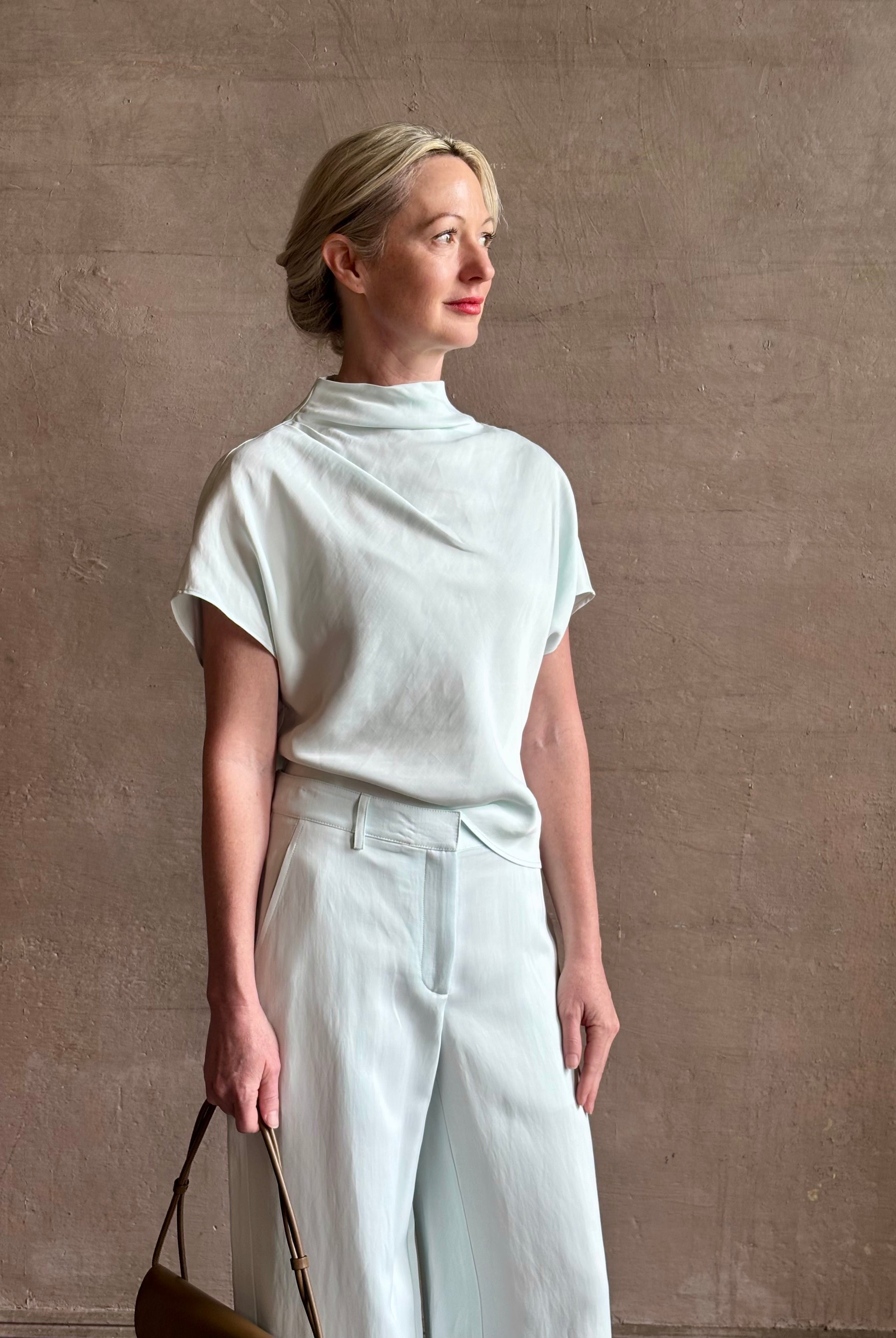 Mature woman wearing refined Mock Neck Tee Blouse from TWP rendered in a subtle sea glass blue hue and crafted from a viscose linen blend made in the USA, with short sleeves, asymmetrical design and quality fabric draping style with matching Dylan pant