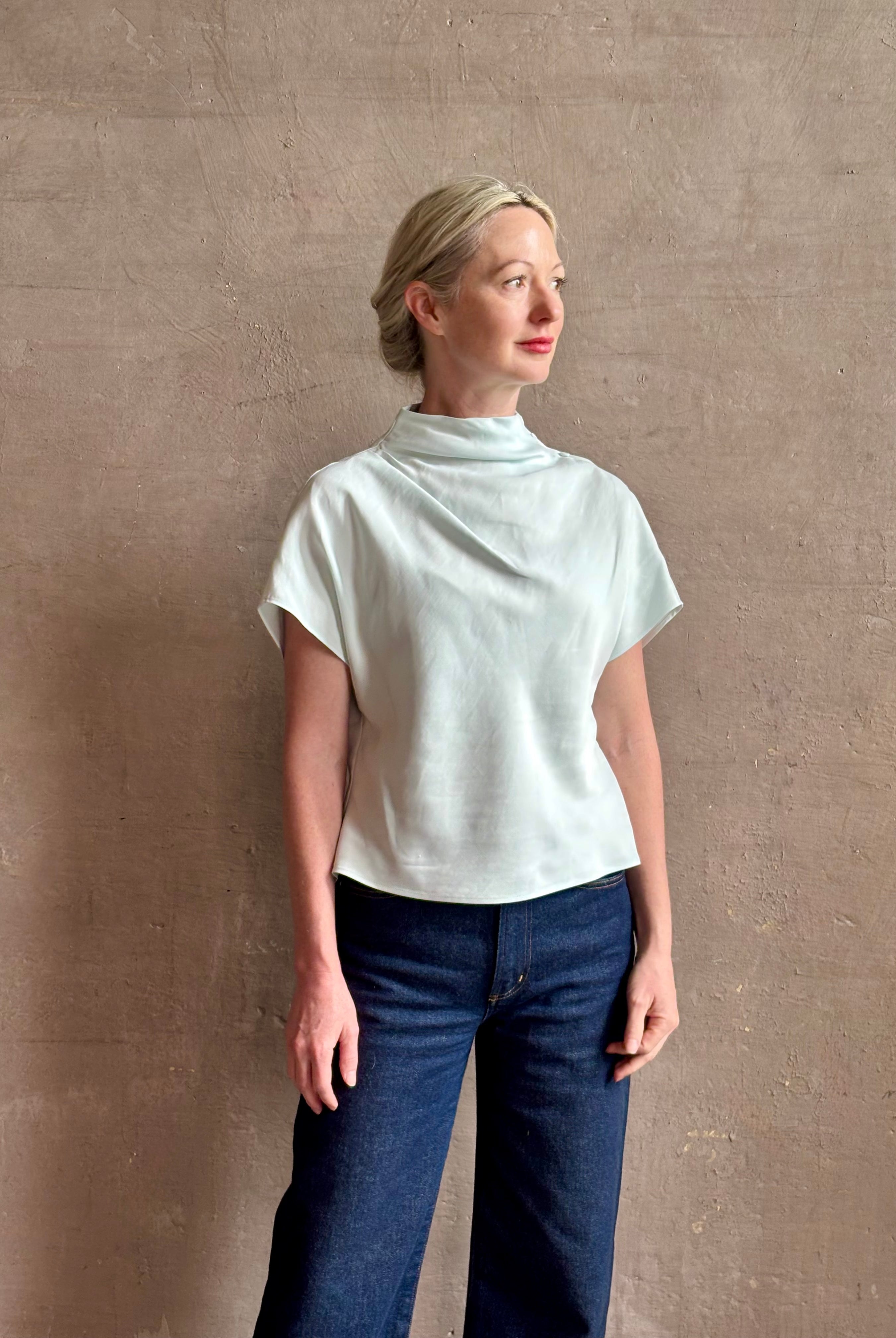 Mature woman wearing refined Mock Neck Tee Blouse from TWP rendered in a subtle sea glass blue hue and crafted from a viscose linen blend made in the USA, with short sleeves, asymmetrical design and quality fabric draping