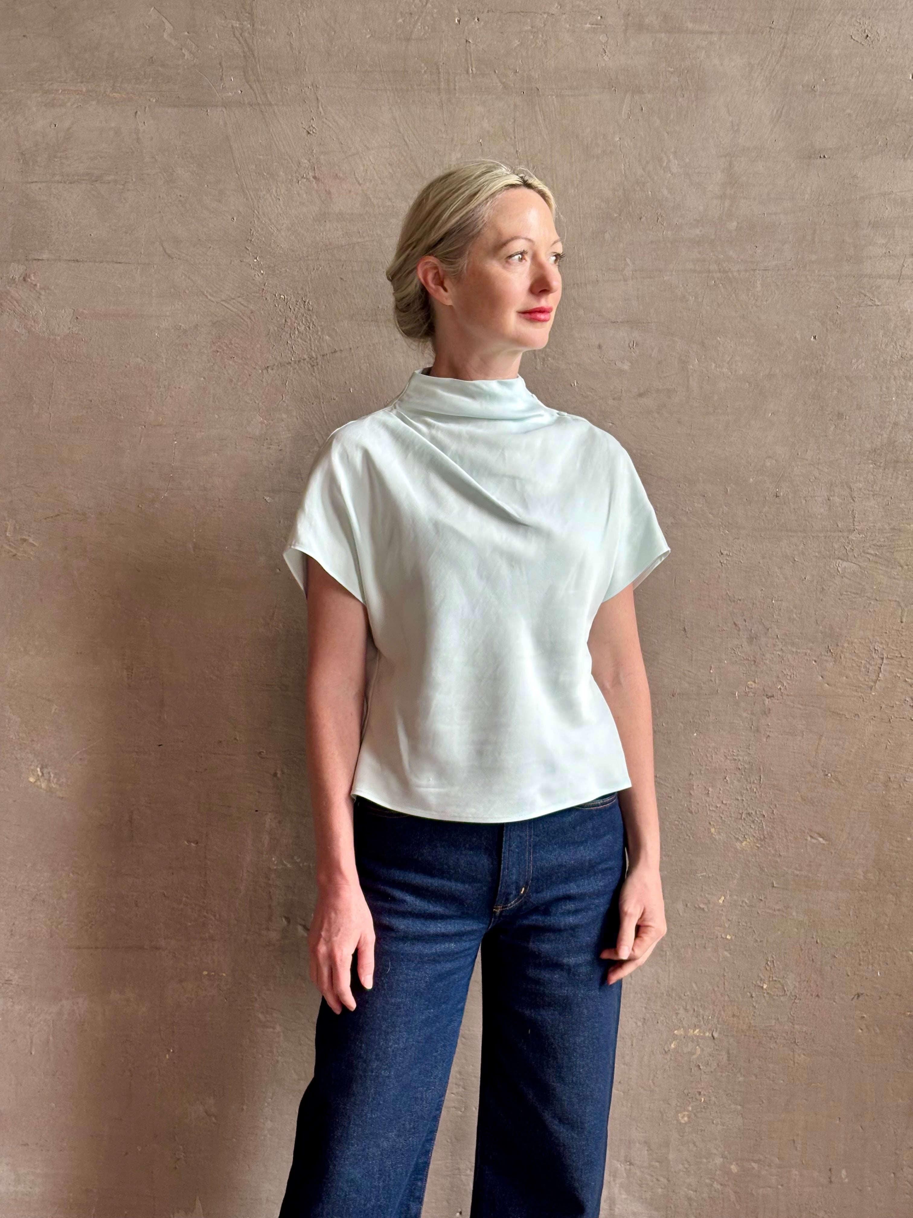 Mature woman wearing refined Mock Neck Tee Blouse from TWP rendered in a subtle sea glass blue hue and crafted from a viscose linen blend made in the USA, with short sleeves, asymmetrical design and quality fabric draping