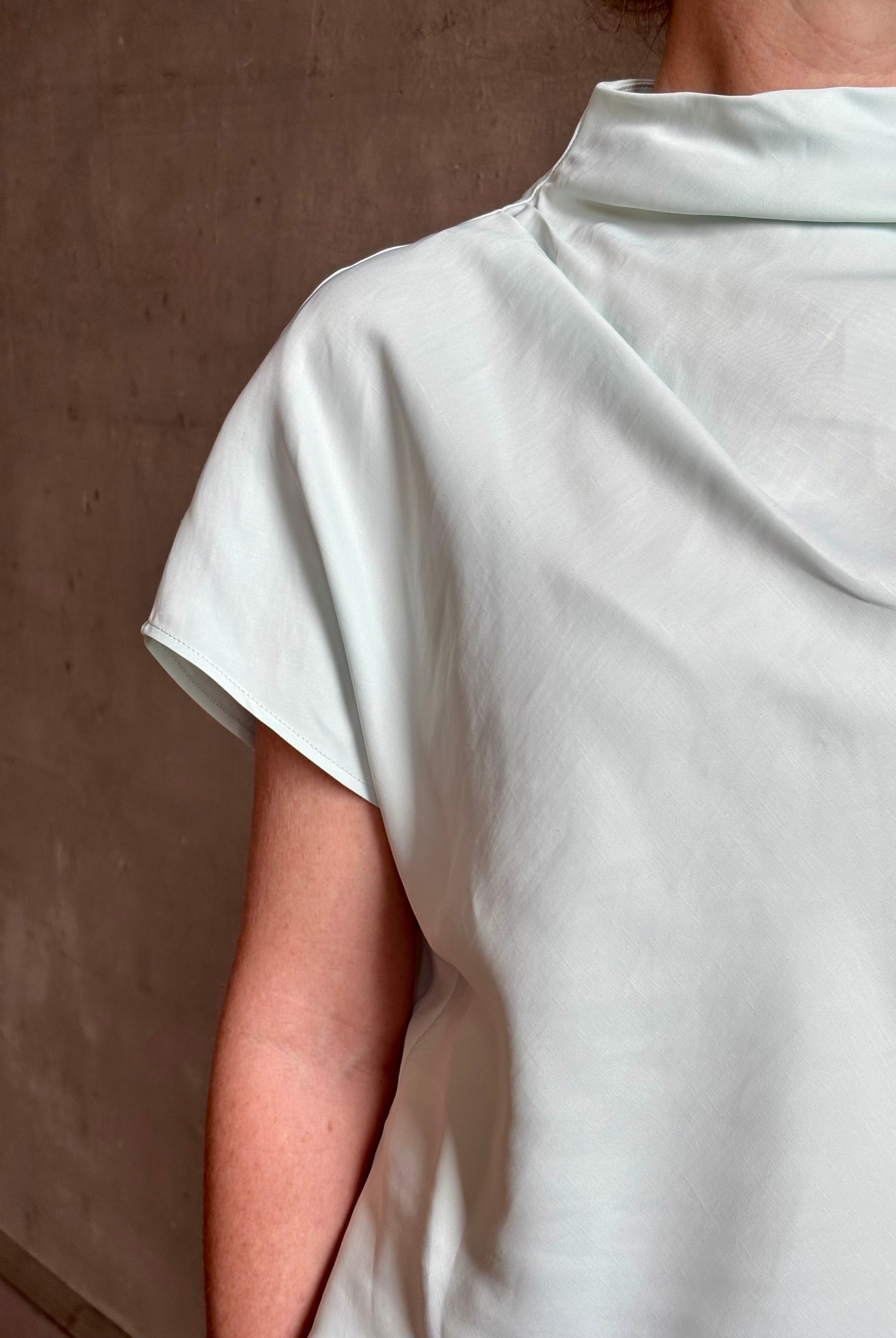 Mature woman wearing refined Mock Neck Tee Blouse from TWP rendered in a subtle sea glass blue hue and crafted from a viscose linen blend made in the USA, with short sleeves, asymmetrical design and quality fabric draping