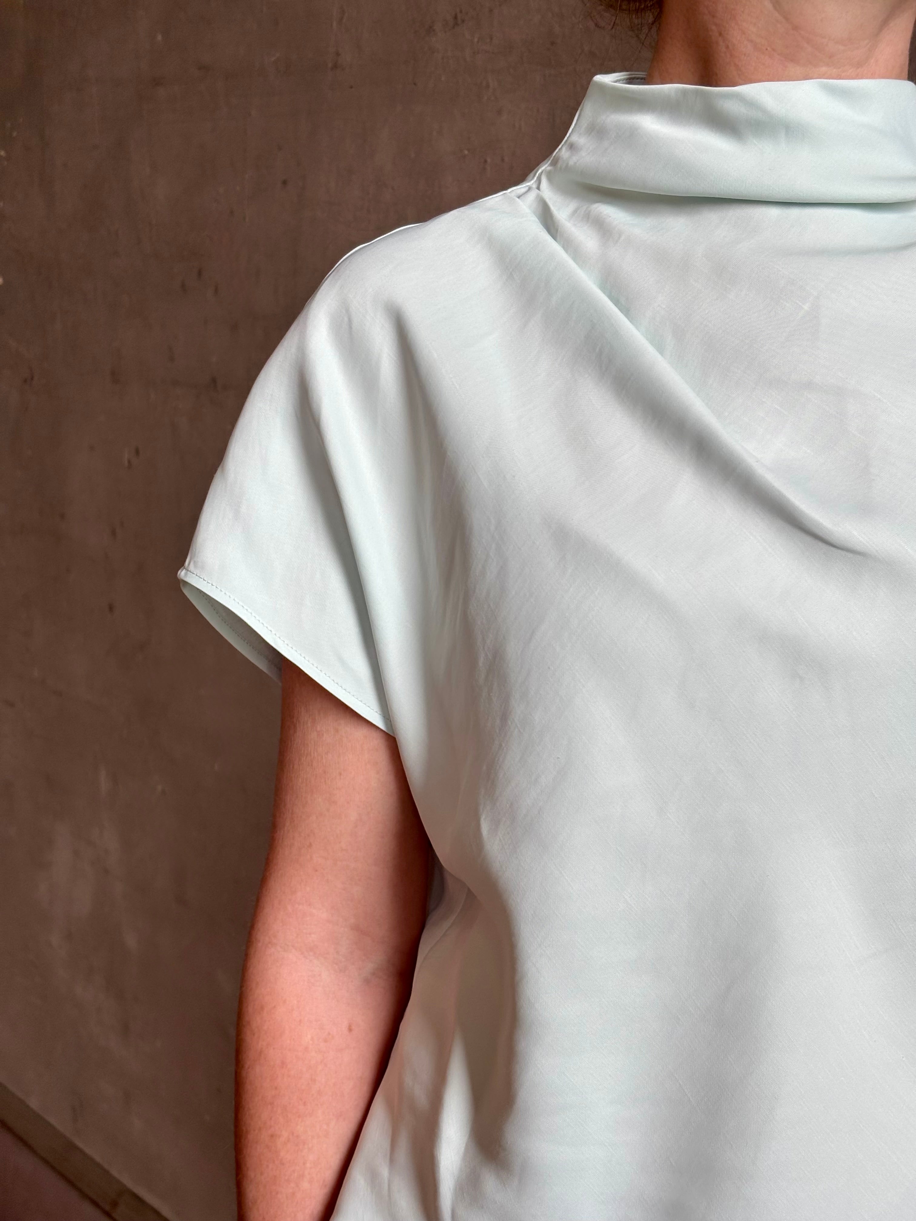 Mature woman wearing refined Mock Neck Tee Blouse from TWP rendered in a subtle sea glass blue hue and crafted from a viscose linen blend made in the USA, with short sleeves, asymmetrical design and quality fabric draping