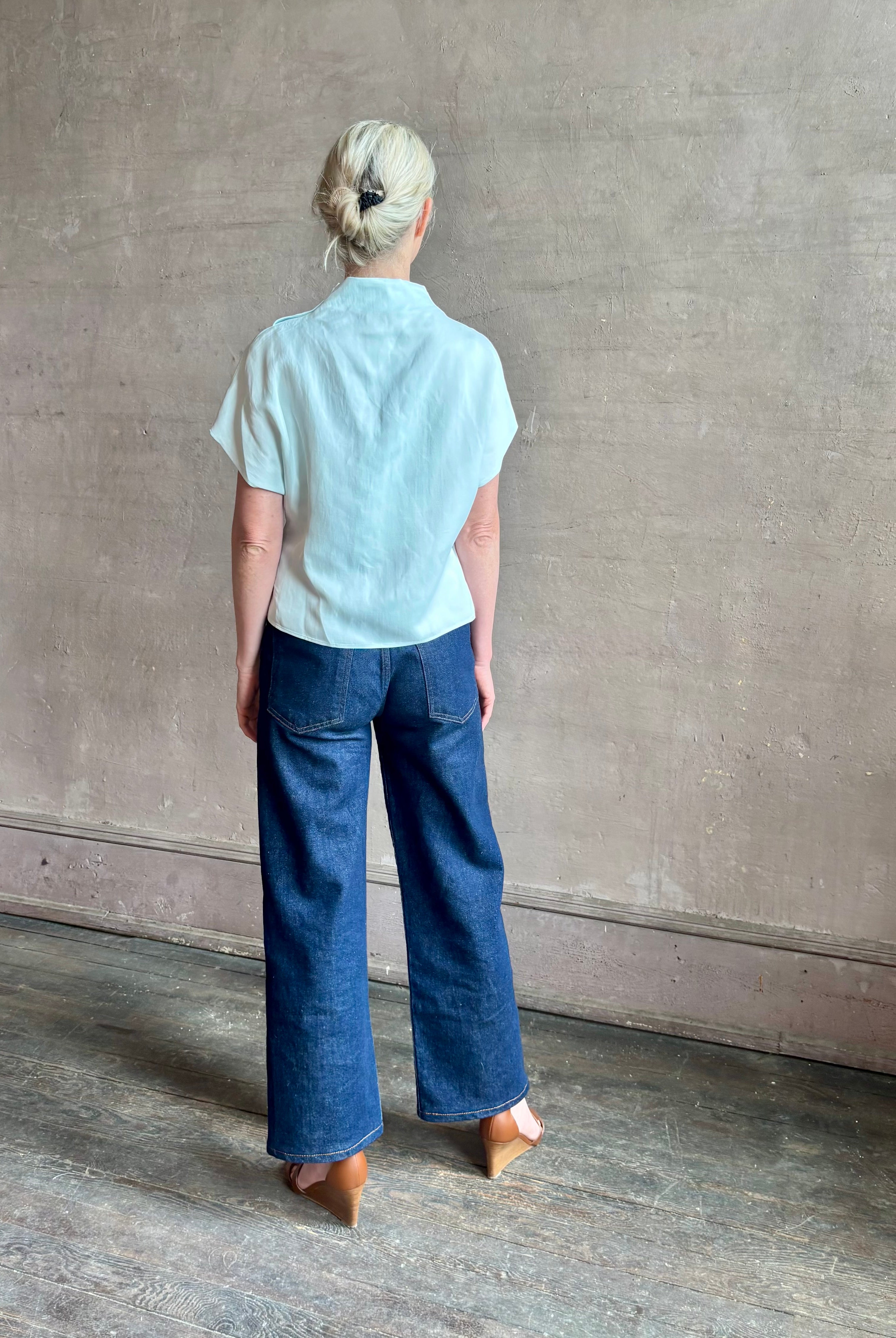 Mature woman wearing refined Mock Neck Tee Blouse from TWP rendered in a subtle sea glass blue hue and crafted from a viscose linen blend made in the USA, with short sleeves, asymmetrical design and quality fabric draping