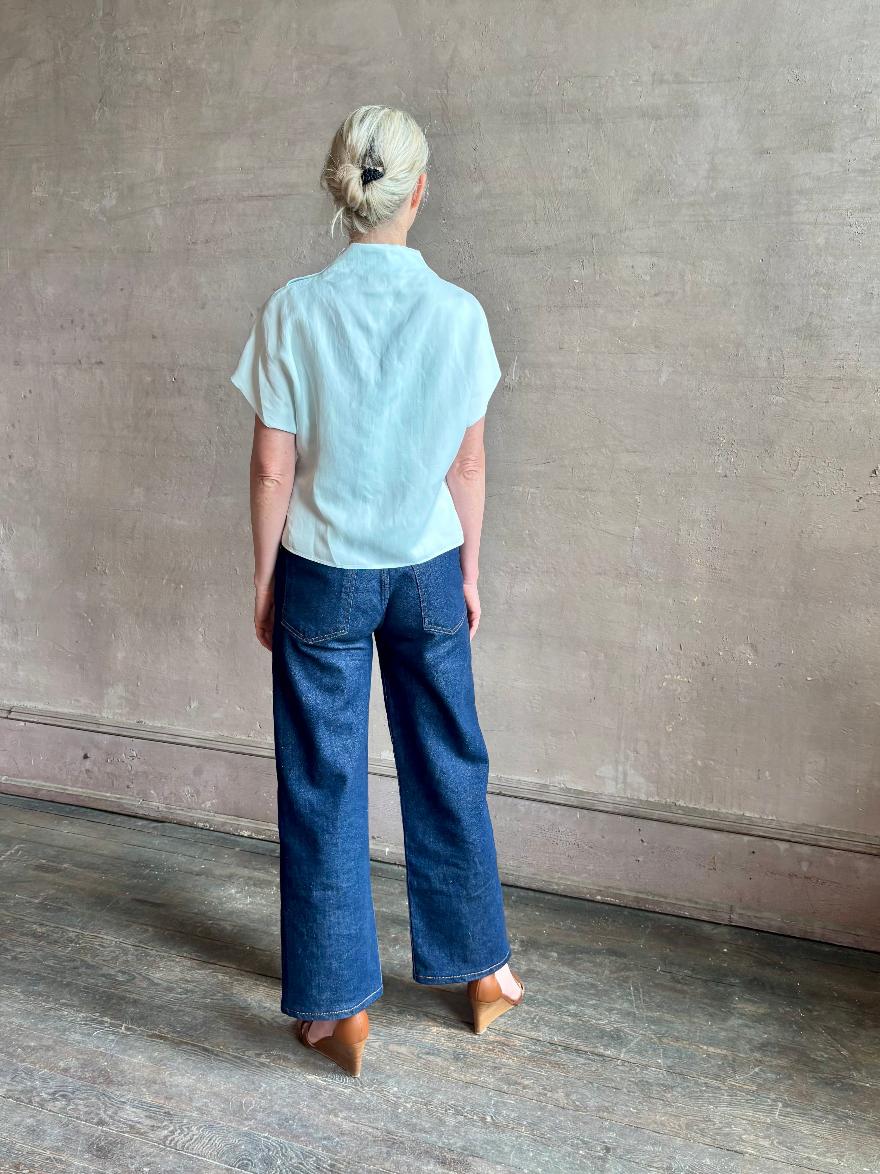 Mature woman wearing refined Mock Neck Tee Blouse from TWP rendered in a subtle sea glass blue hue and crafted from a viscose linen blend made in the USA, with short sleeves, asymmetrical design and quality fabric draping