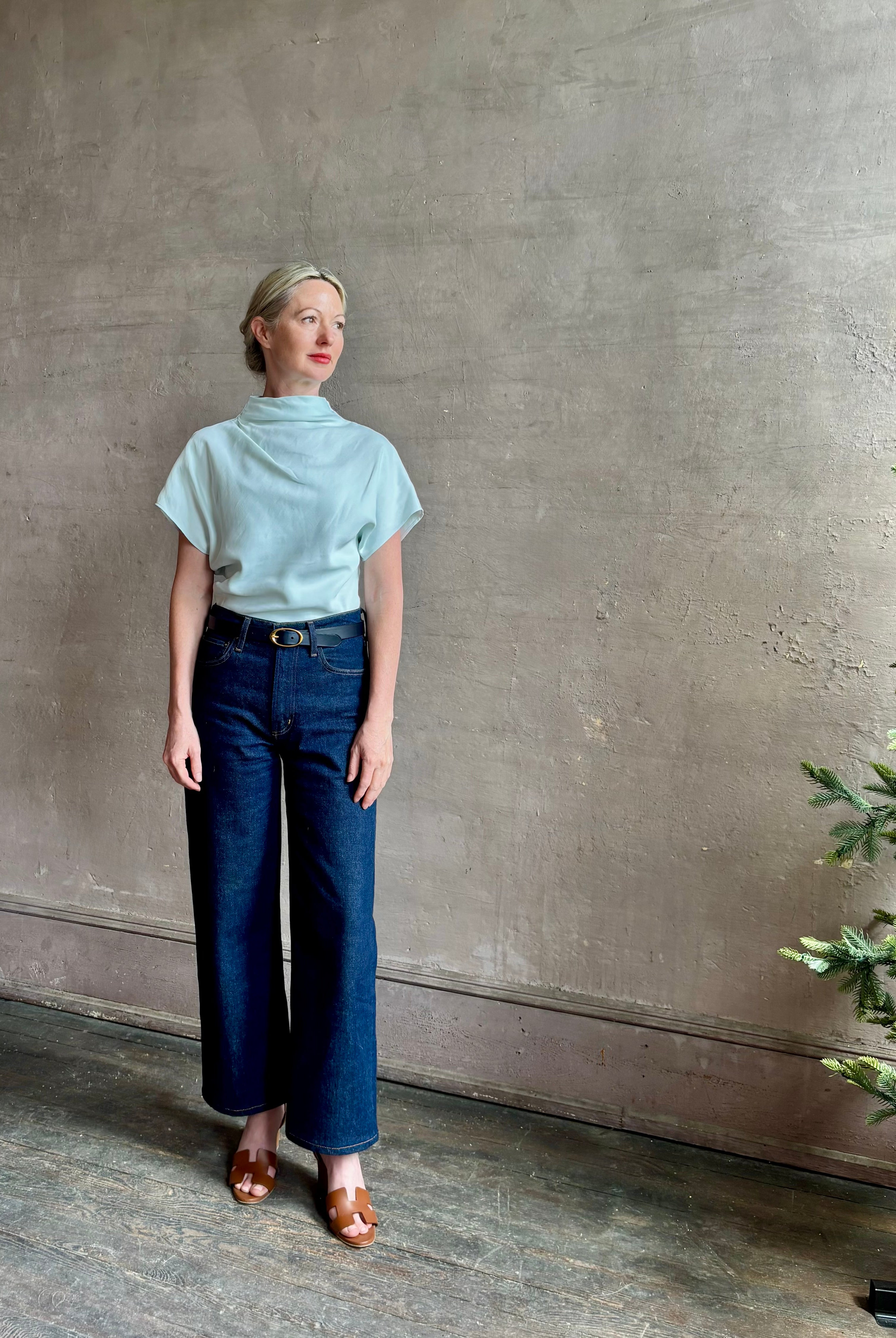 Mature woman wearing refined Mock Neck Tee Blouse from TWP rendered in a subtle sea glass blue hue and crafted from a viscose linen blend made in the USA, with short sleeves, asymmetrical design and quality fabric draping
