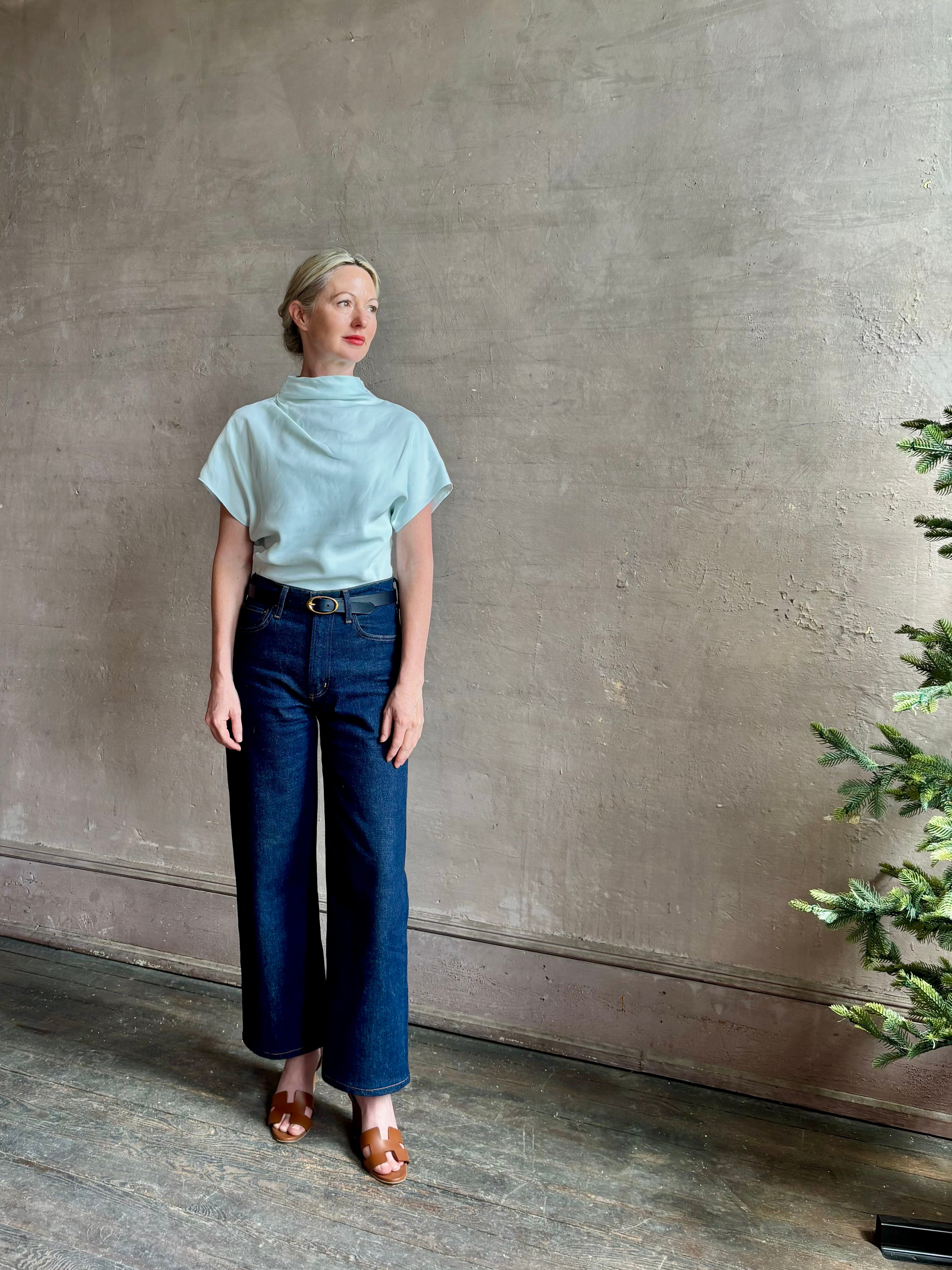 Mature woman wearing refined Mock Neck Tee Blouse from TWP rendered in a subtle sea glass blue hue and crafted from a viscose linen blend made in the USA, with short sleeves, asymmetrical design and quality fabric draping