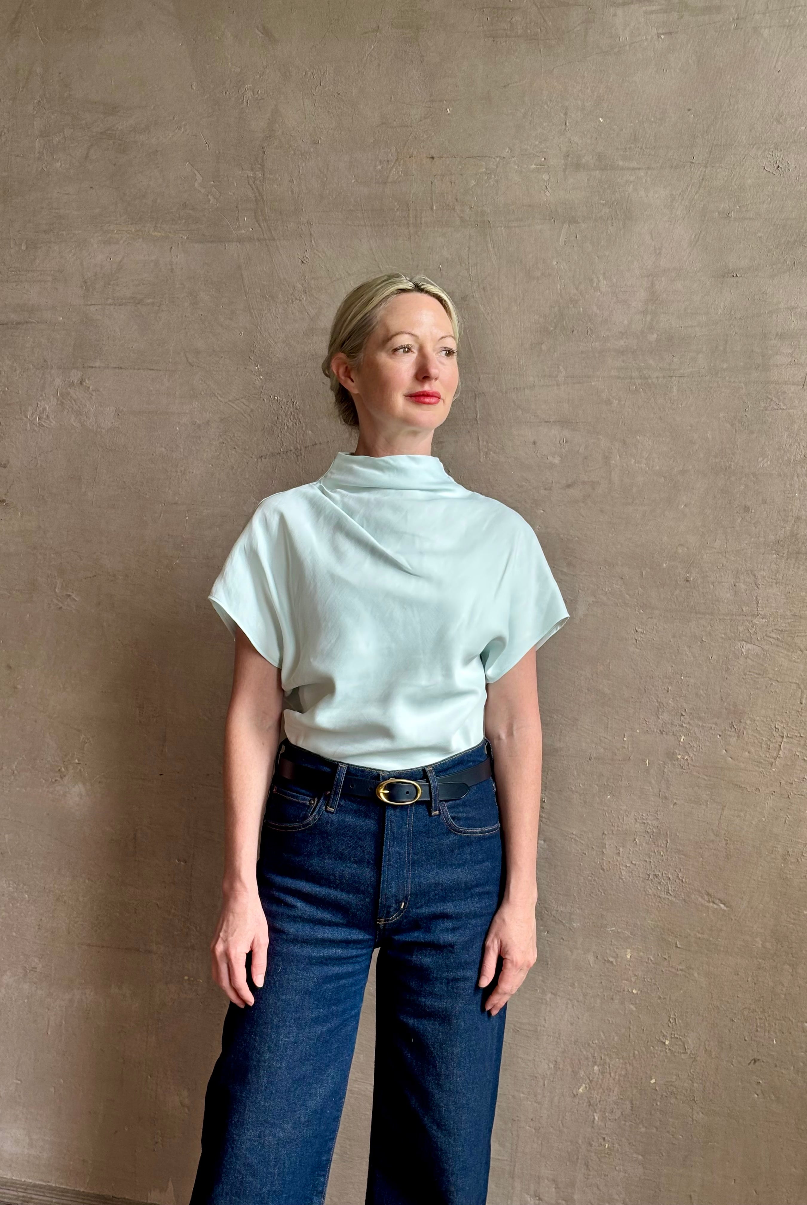 Mature woman wearing refined Mock Neck Tee Blouse from TWP rendered in a subtle sea glass blue hue and crafted from a viscose linen blend made in the USA, with short sleeves, asymmetrical design and quality fabric draping