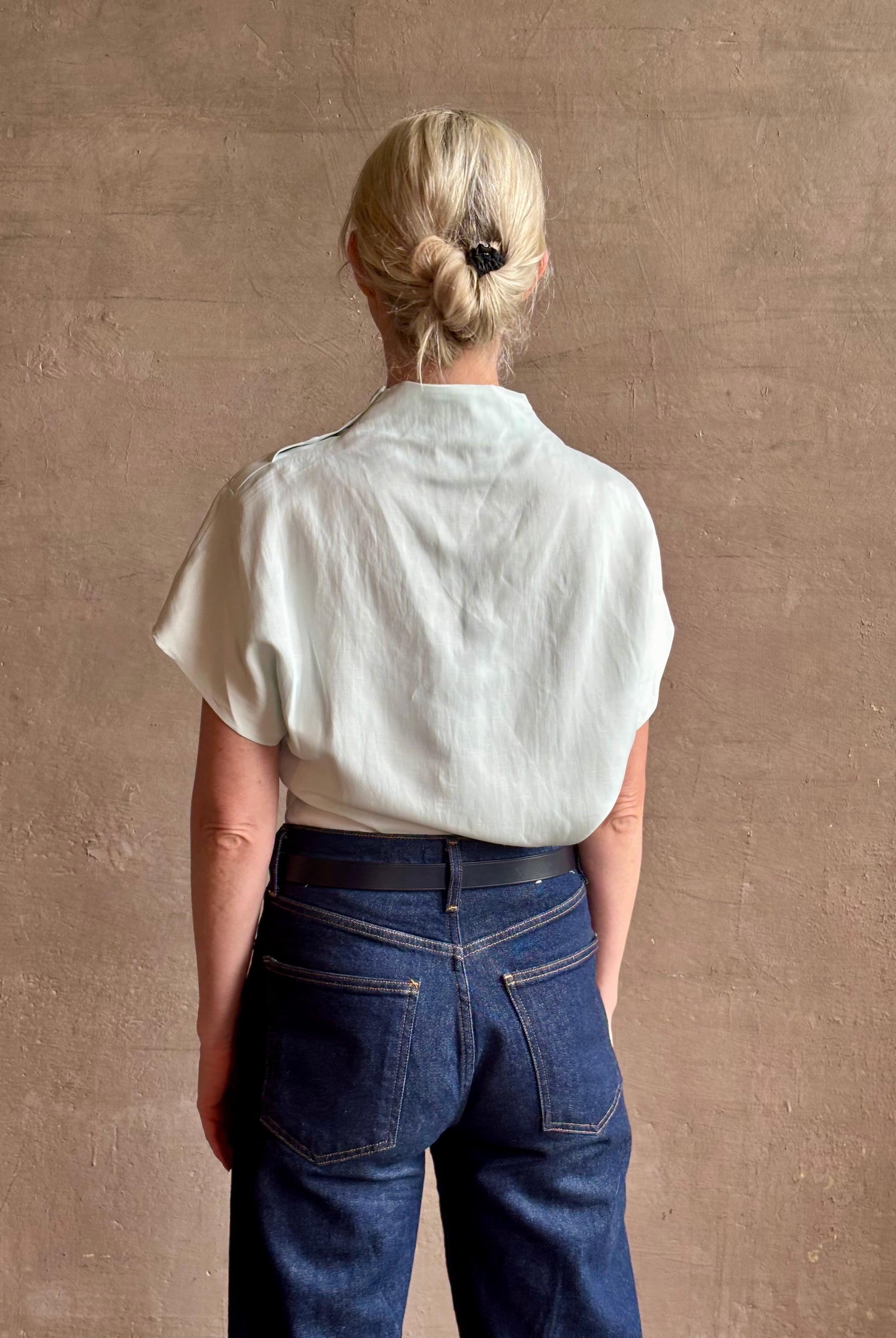 Mature woman wearing refined Mock Neck Tee Blouse from TWP rendered in a subtle sea glass blue hue and crafted from a viscose linen blend made in the USA, with short sleeves, asymmetrical design and quality fabric draping