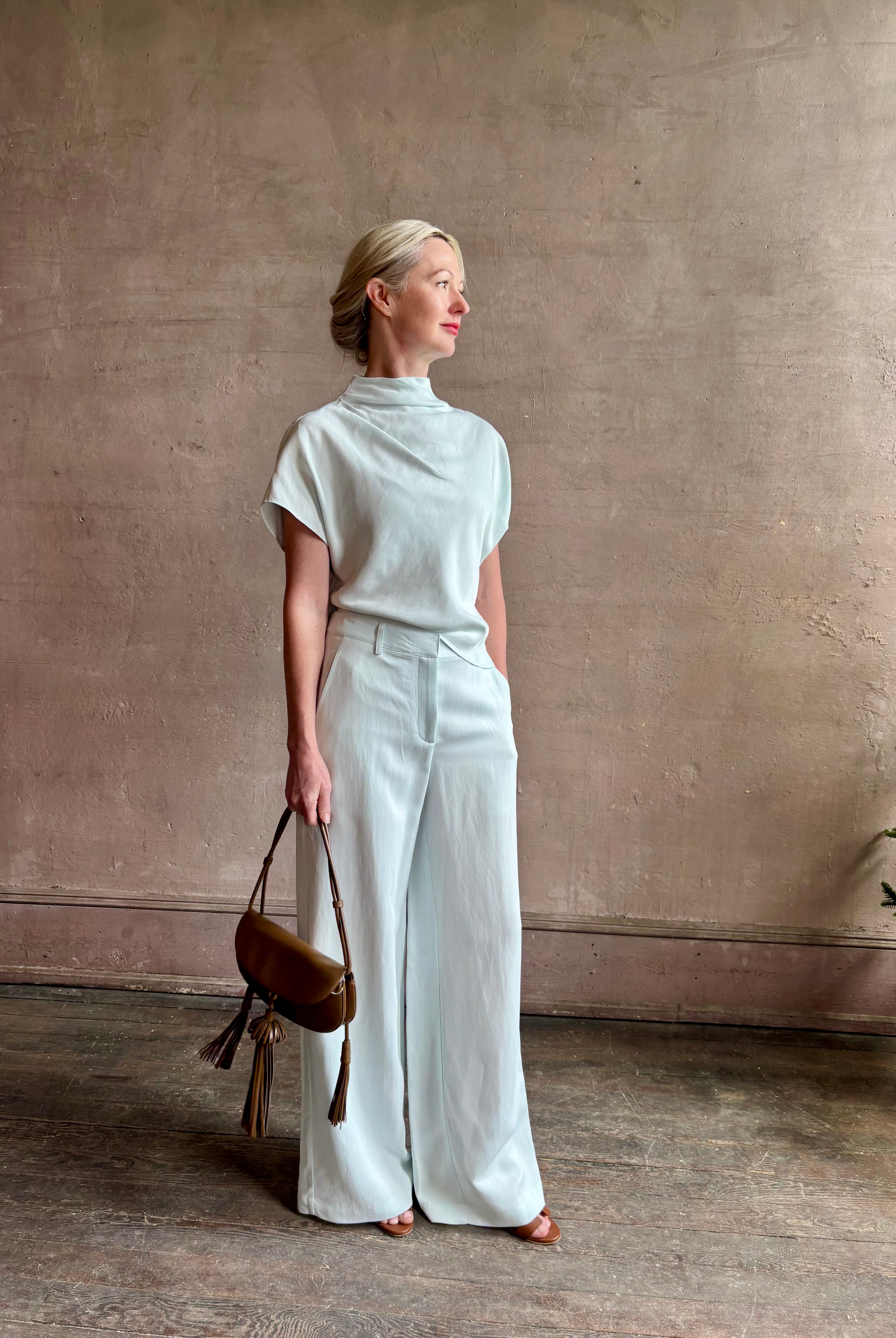 Mature woman wearing refined Mock Neck Tee Blouse from TWP rendered in a subtle sea glass blue hue and crafted from a viscose linen blend made in the USA, with short sleeves, asymmetrical design and quality fabric draping