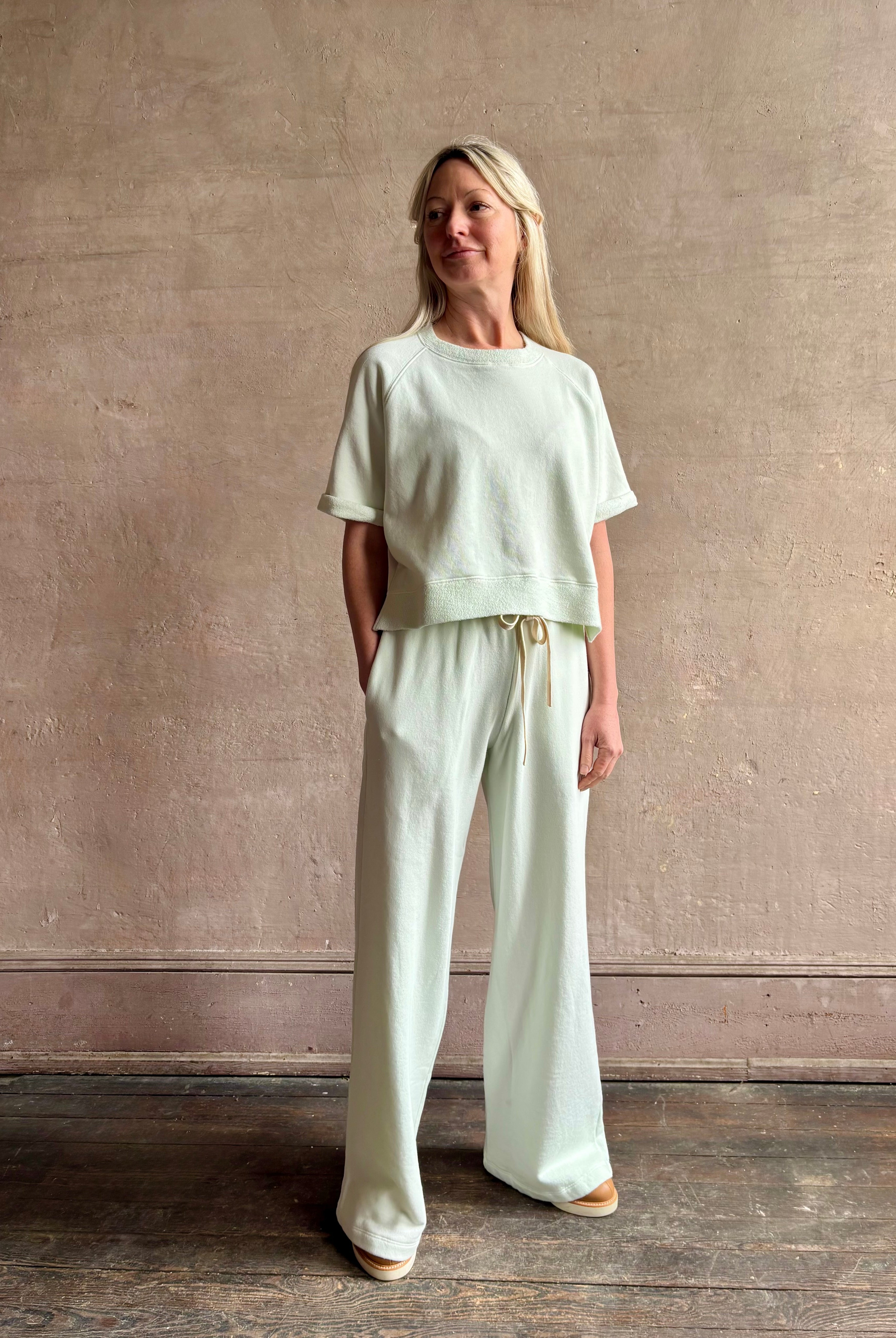 Mature woman wearing Slouchy Rolled Tee Top from TWP rendered in a sea glass hue and crafted from cotton with raglan short sleeves, crew neck and relaxed silhouette styled with matching PJ pant for the complete luxury lounge set