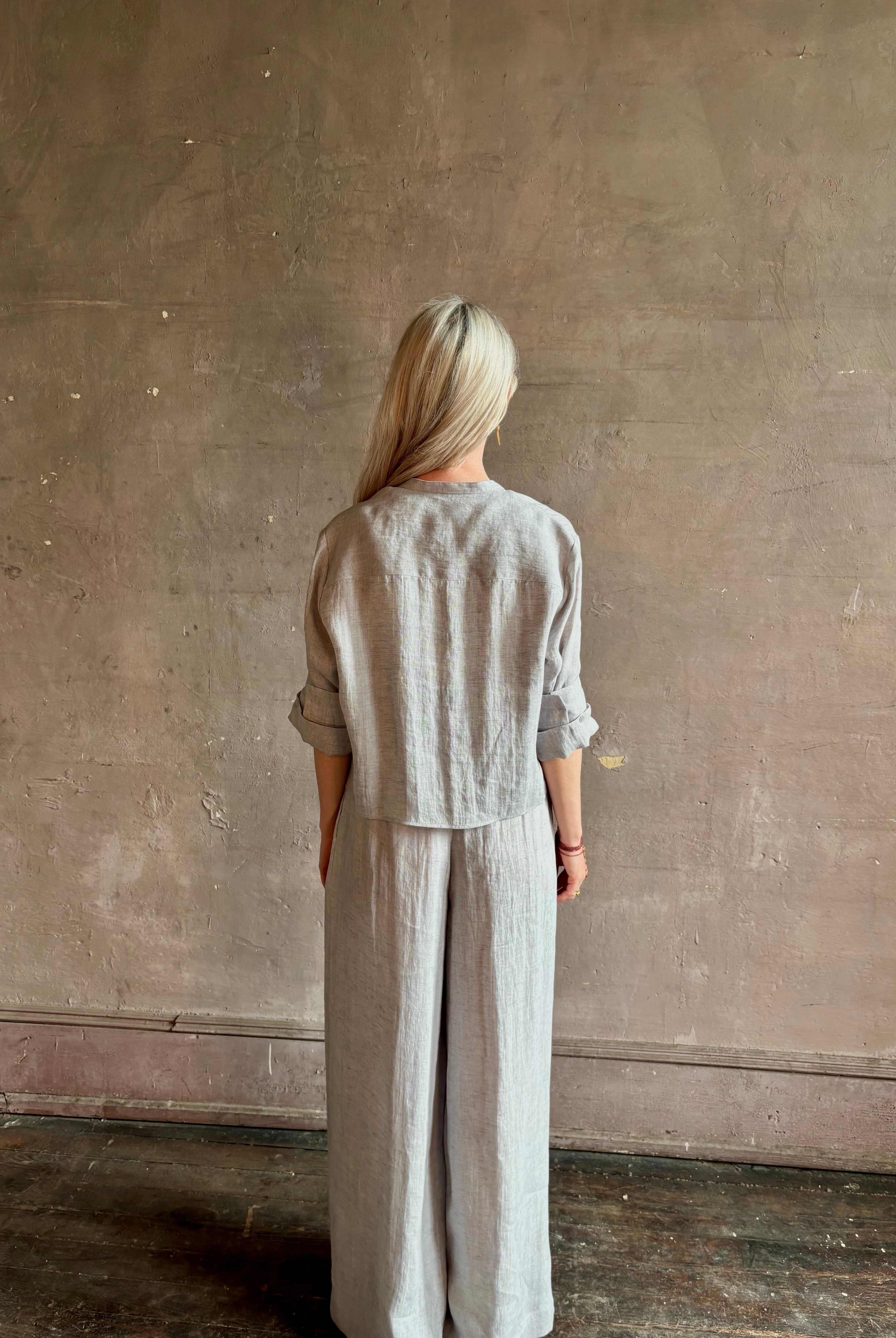 Mature woman wearing Stand Collar Cut Off Top from TWP rendered in a versatile light heather grey and crafted from 100% hemp with round mandarin collar, button front, half sleeves and made in the USA