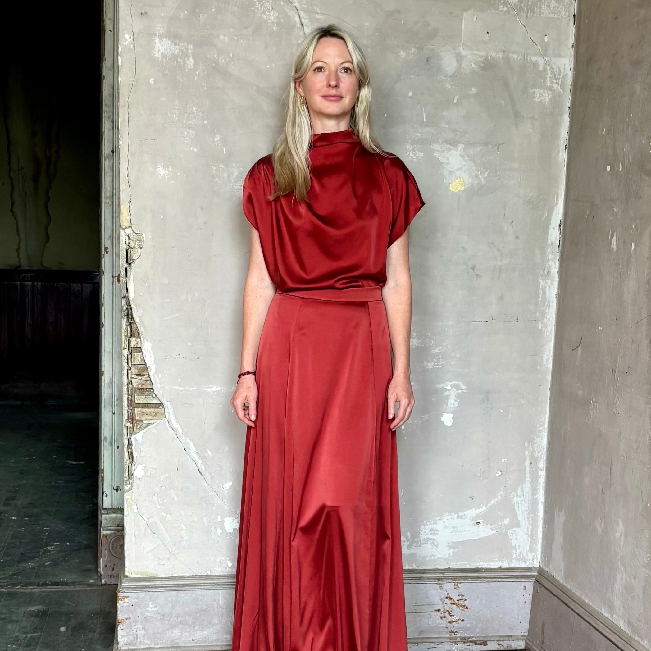 Mature woman wearing Wish You Would blouse from TWP rendered in a rich currant red and crafted from a luxurious silk blend with a mock neck, asymetrical design and draping short sleeves shoulder button closure and matching skirt
