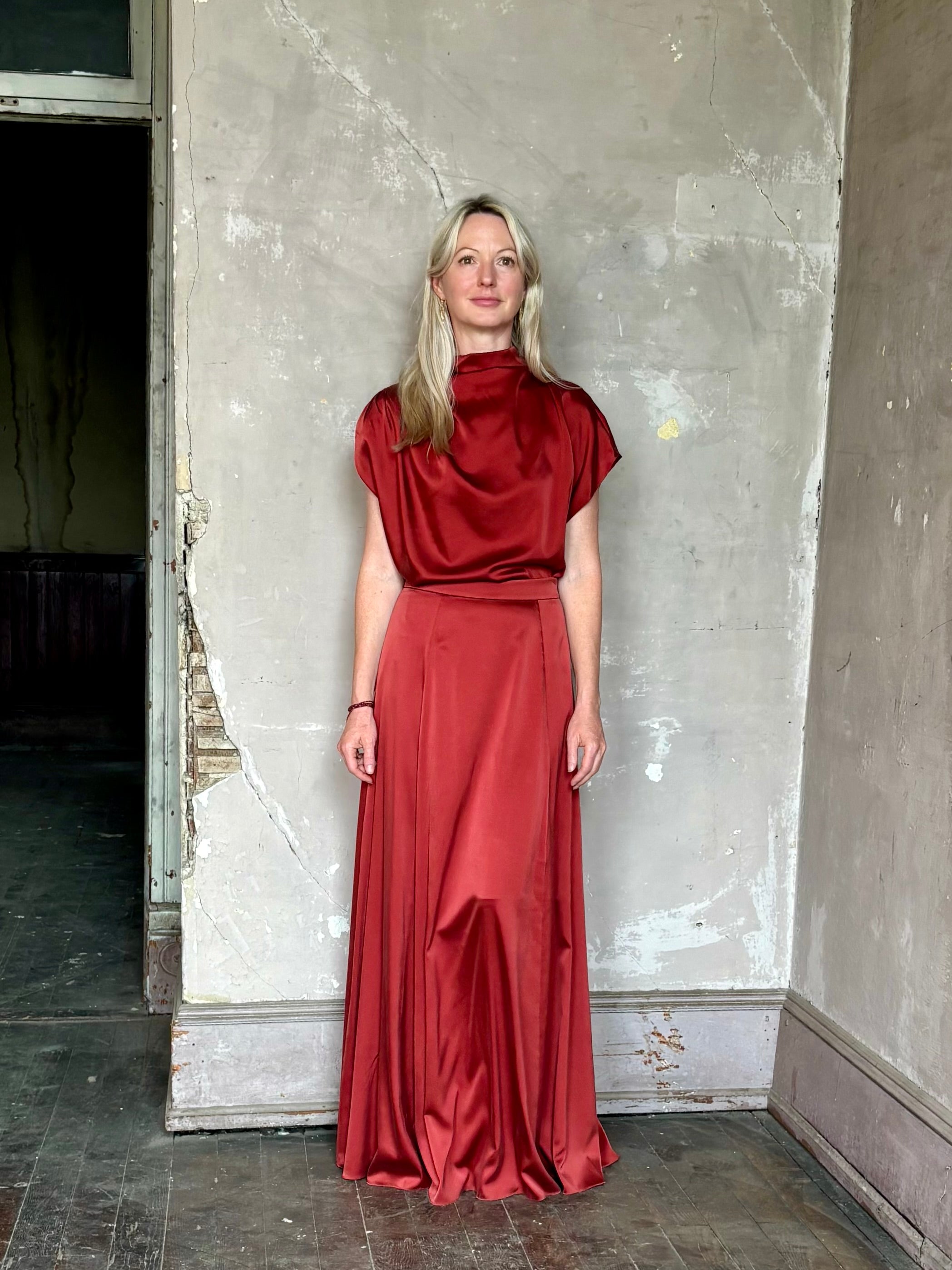Mature woman wearing Wish You Would blouse from TWP rendered in a rich currant red and crafted from a luxurious silk blend with a mock neck, asymetrical design and draping short sleeves shoulder button closure and matching skirt