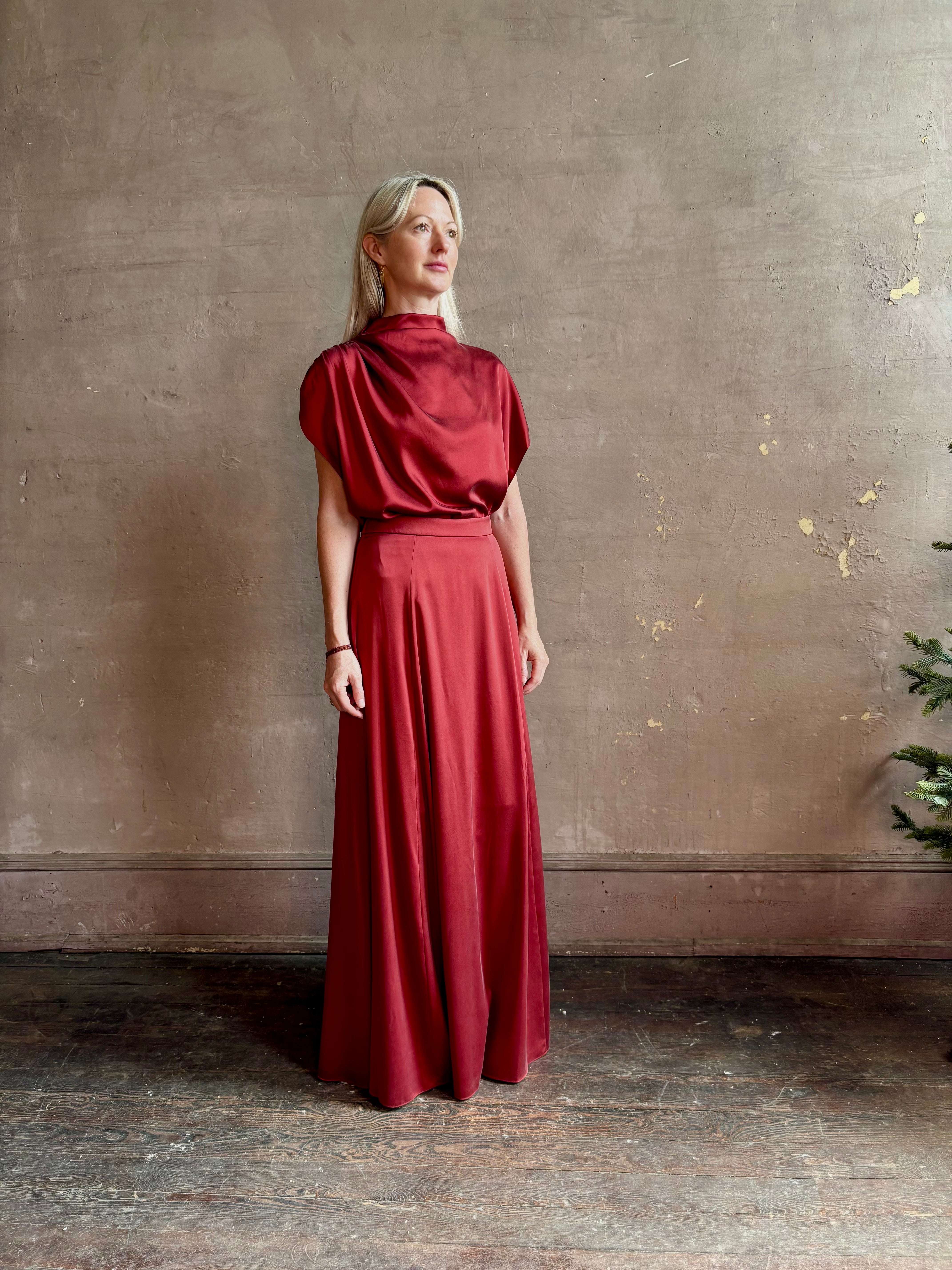 Mature woman wearing Wish You Would blouse from TWP rendered in a rich currant red and crafted from a luxurious silk blend with a mock neck, asymetrical design and draping short sleeves shoulder button closure and matching skirt