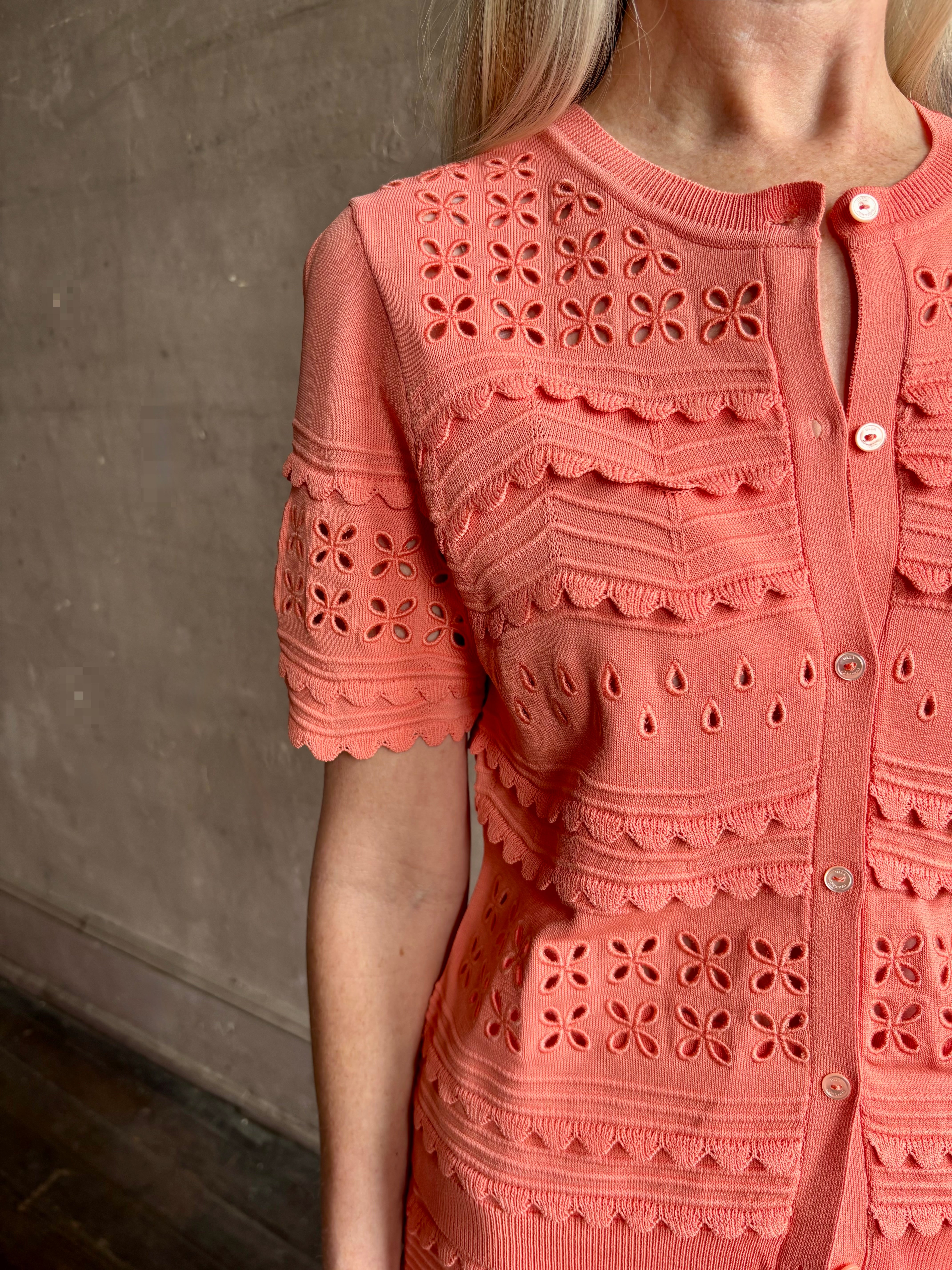 Woman wearing the romantic Abriana Eyelet Knit Cardigan from Ulla Johnson rendered in a luminous coral hue and crafted from luxury knit with round neck, button front and short sleeves