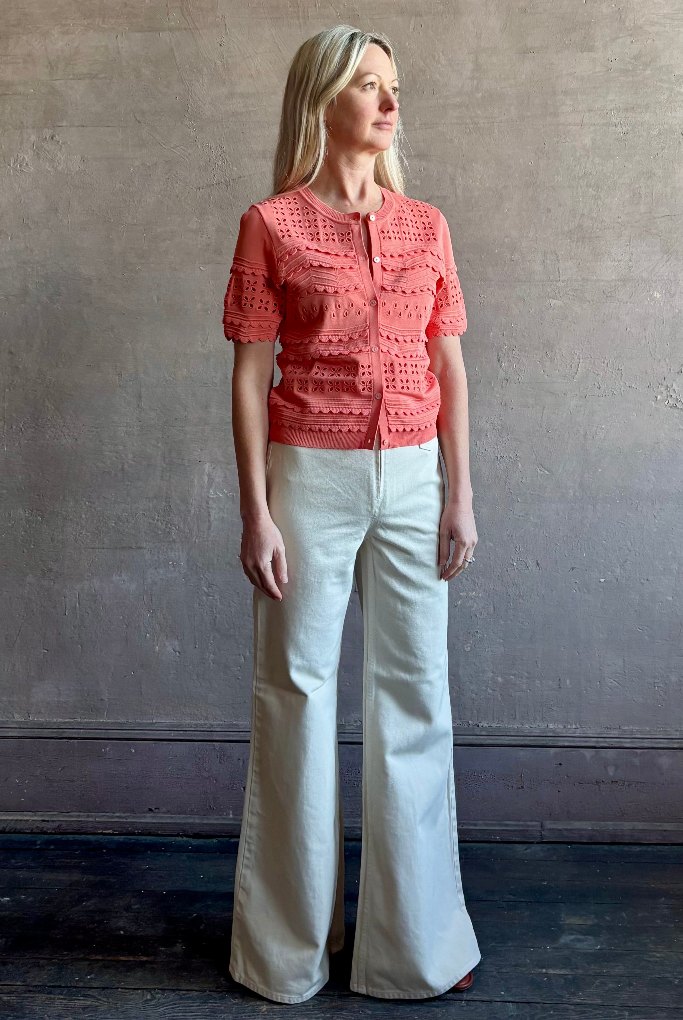 Woman wearing the romantic Abriana Eyelet Knit Cardigan from Ulla Johnson rendered in a luminous coral hue and crafted from luxury knit with round neck, button front and short sleeves