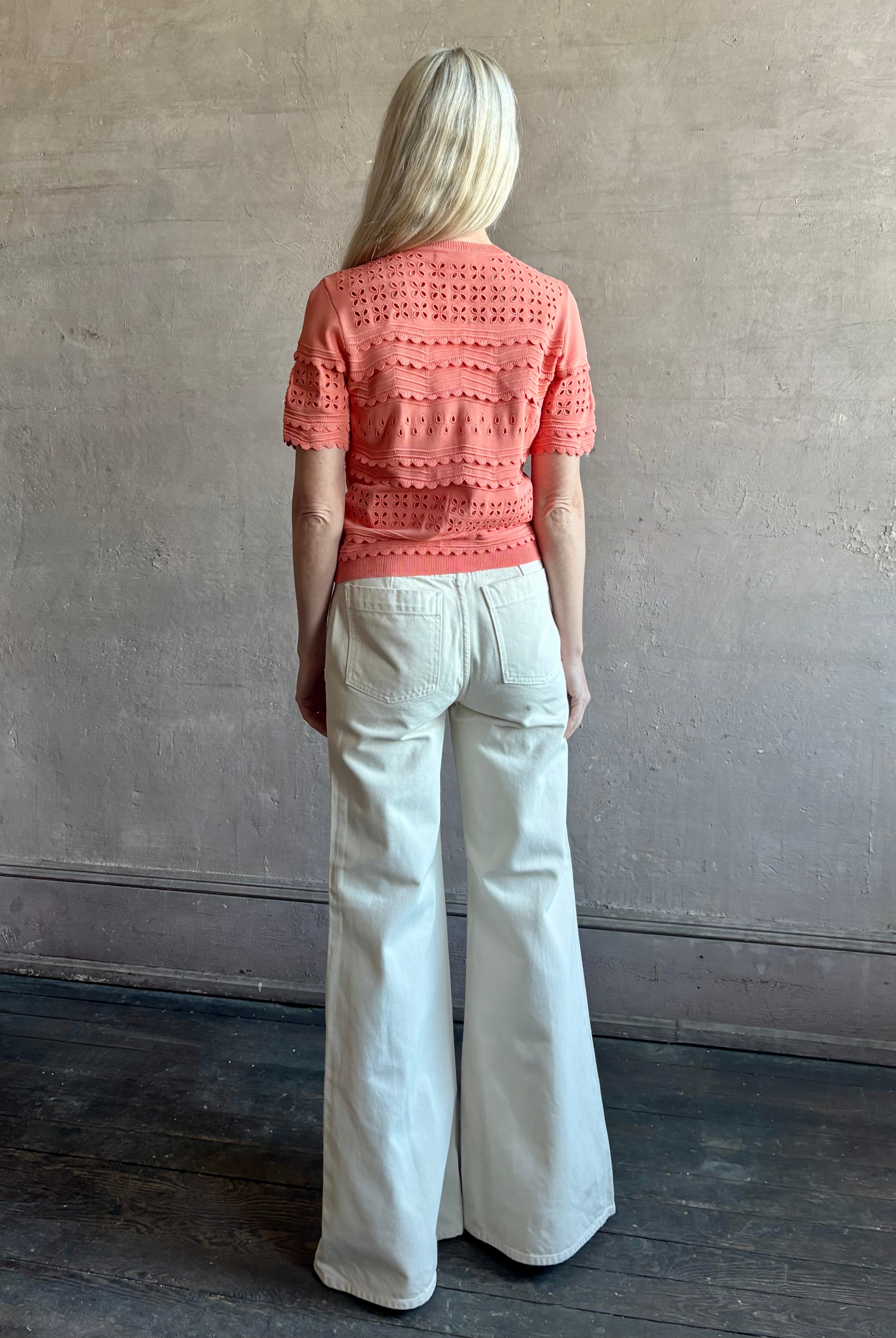 Woman wearing the romantic Abriana Eyelet Knit Cardigan from Ulla Johnson rendered in a luminous coral hue and crafted from luxury knit with round neck, button front and short sleeves