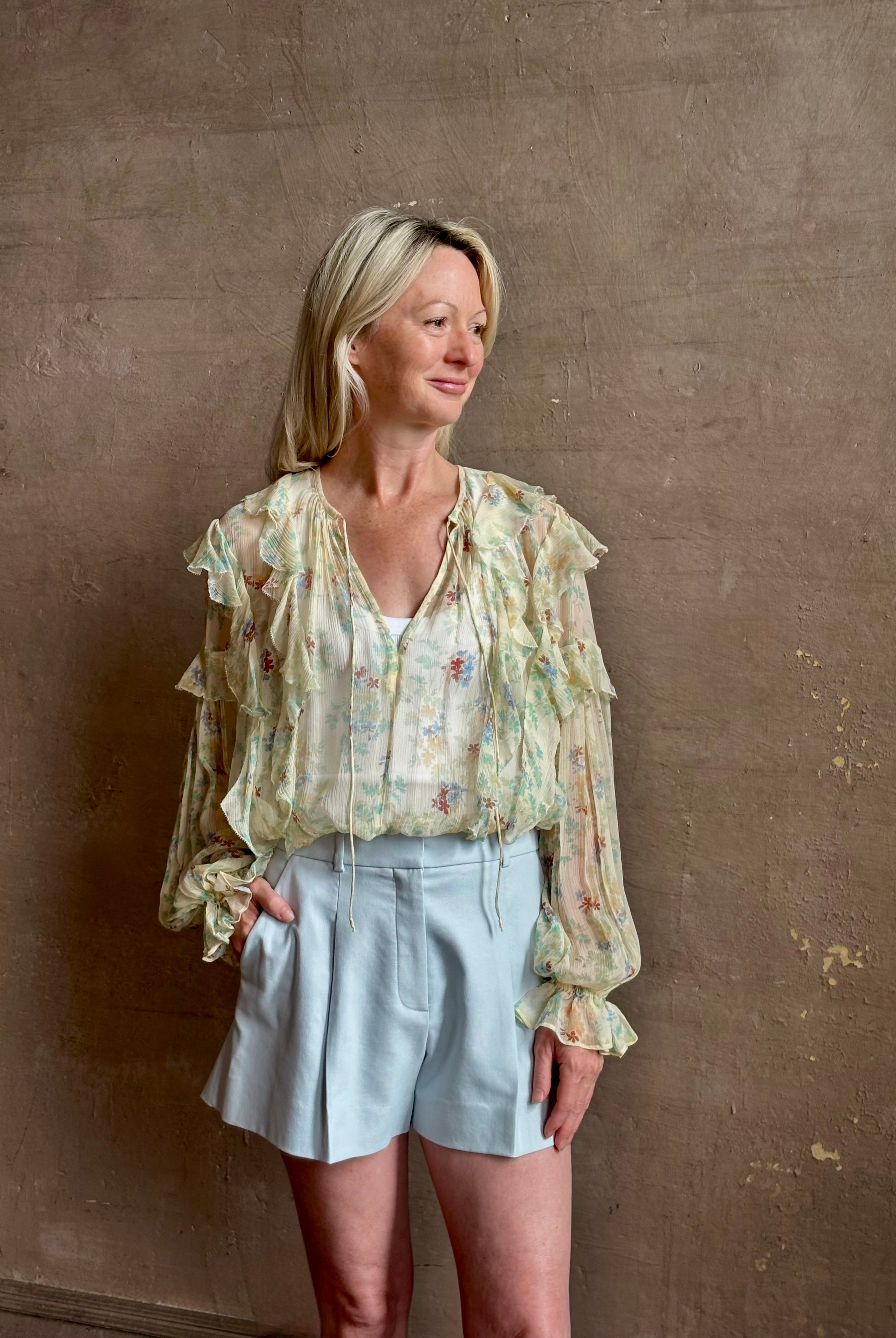 Mature woman wearing Delfina Long Sleeve Silk Blouse from Ulla Johnson rendered in a light yellow body adorned with a delicate, scattered tiny floral print with v-neck, drawstring waist, ruffle lined sleeves and linear ruffle details at front and back