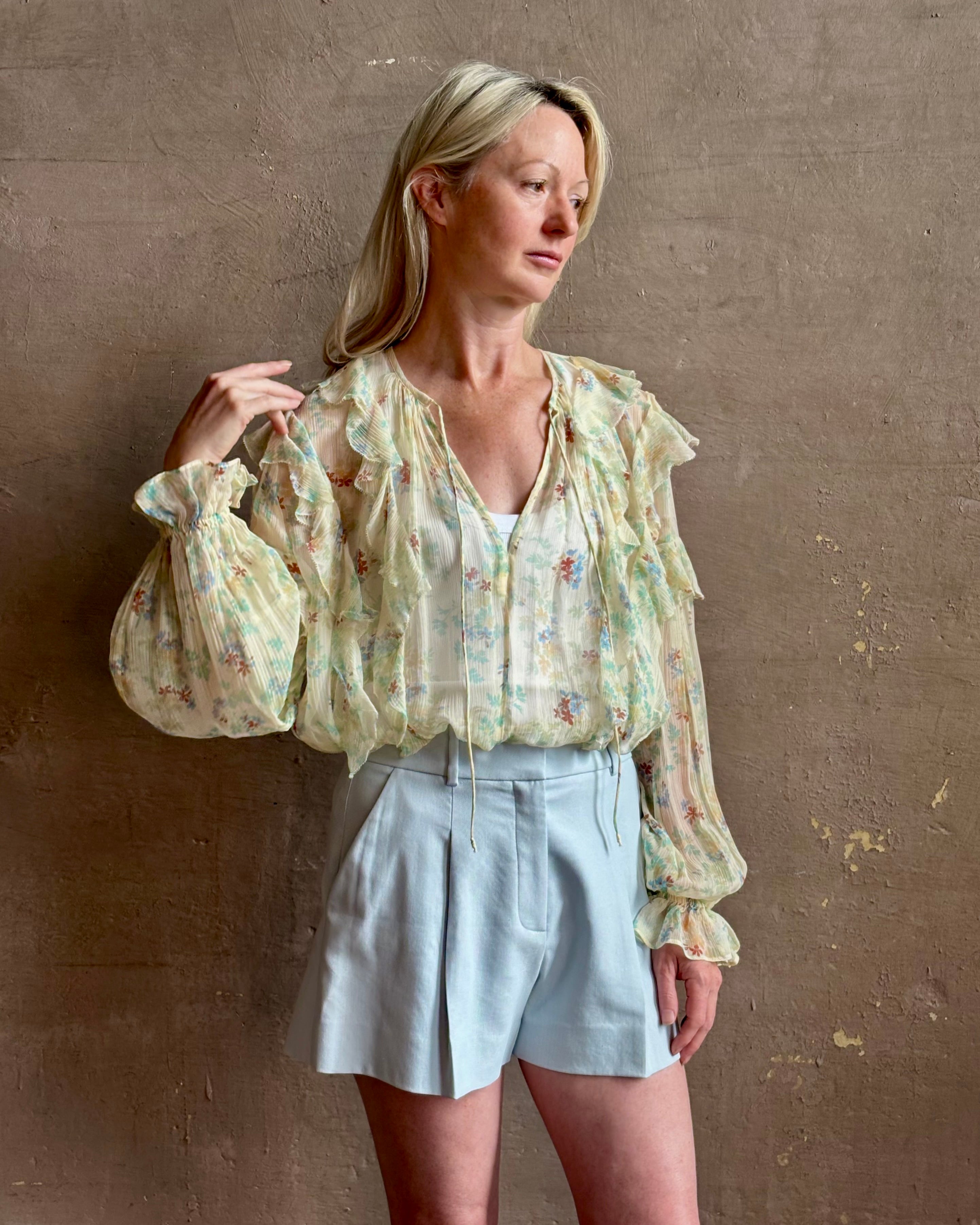 Mature woman wearing Delfina Long Sleeve Silk Blouse from Ulla Johnson rendered in a light yellow body adorned with a delicate, scattered tiny floral print with v-neck, drawstring waist, ruffle lined sleeves and linear ruffle details at front and back