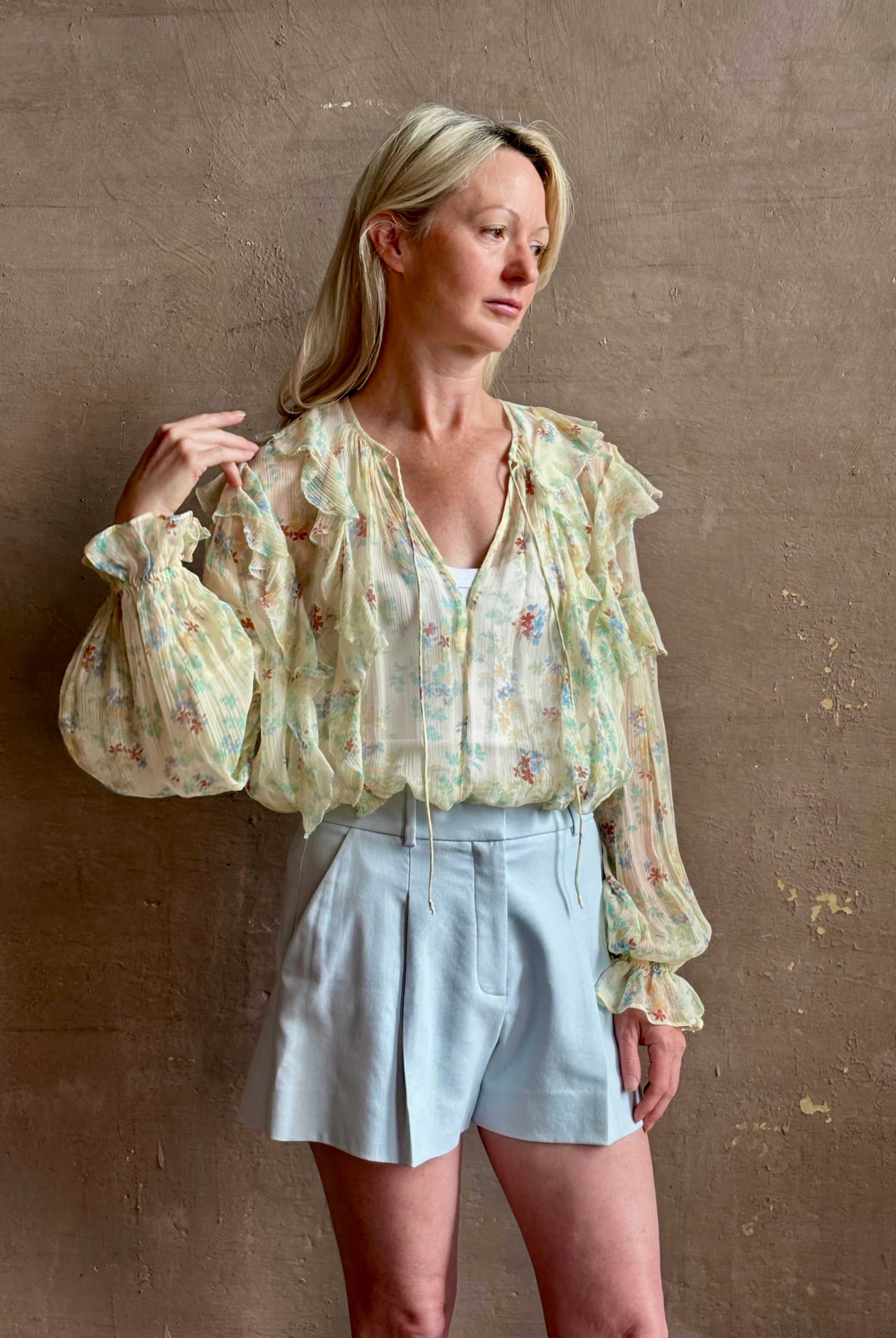 Mature woman wearing Delfina Long Sleeve Silk Blouse from Ulla Johnson rendered in a light yellow body adorned with a delicate, scattered tiny floral print with v-neck, drawstring waist, ruffle lined sleeves and linear ruffle details at front and back