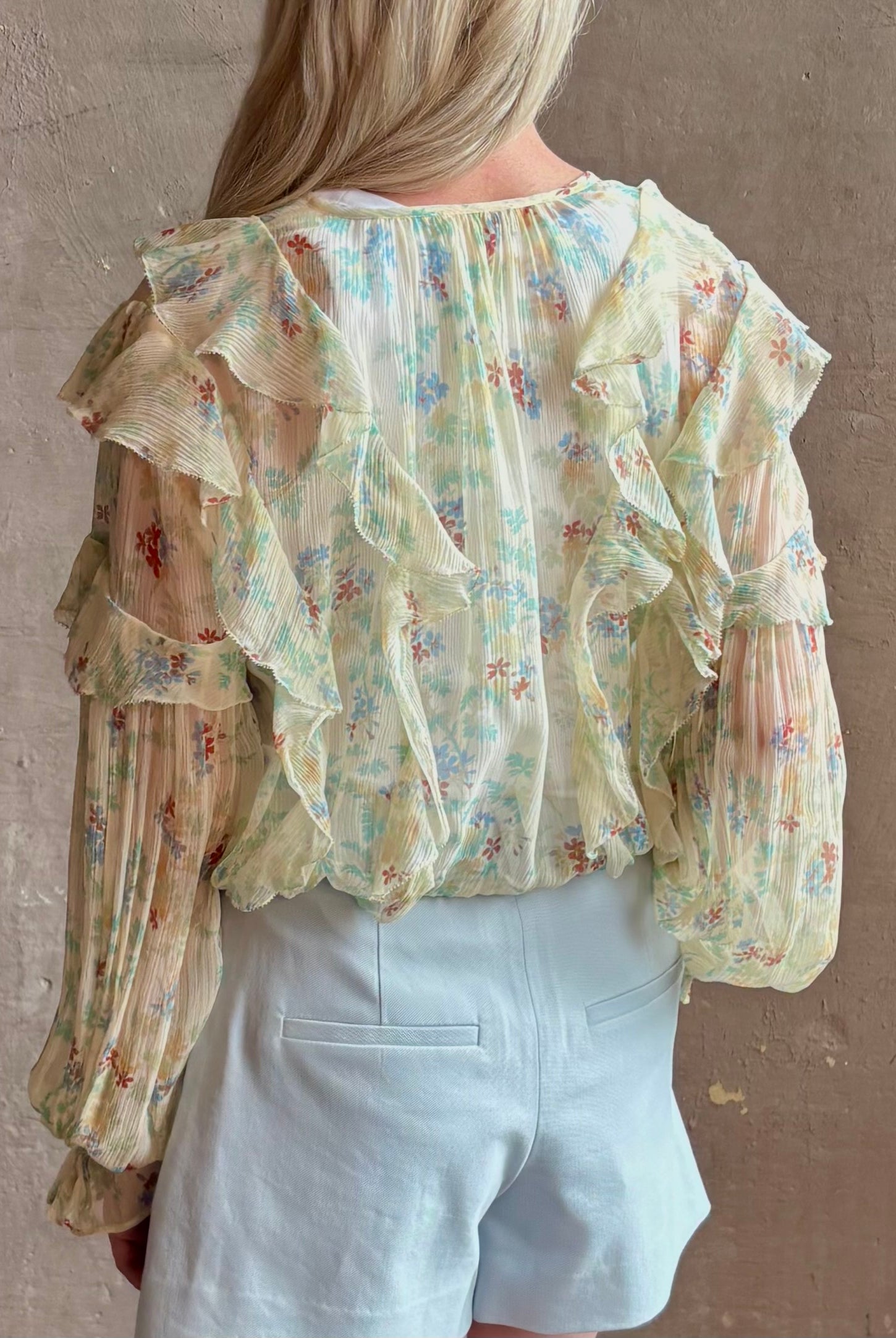 Mature woman wearing Delfina Long Sleeve Silk Blouse from Ulla Johnson rendered in a light yellow body adorned with a delicate, scattered tiny floral print with v-neck, drawstring waist, ruffle lined sleeves and linear ruffle details at front and back