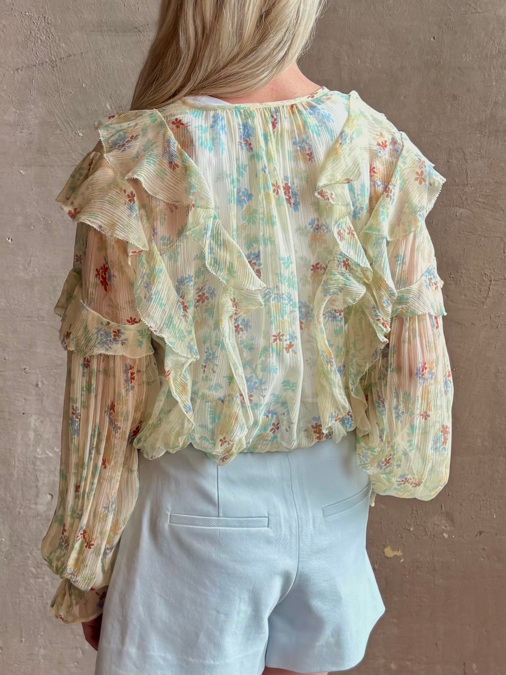 Mature woman wearing Delfina Long Sleeve Silk Blouse from Ulla Johnson rendered in a light yellow body adorned with a delicate, scattered tiny floral print with v-neck, drawstring waist, ruffle lined sleeves and linear ruffle details at front and back