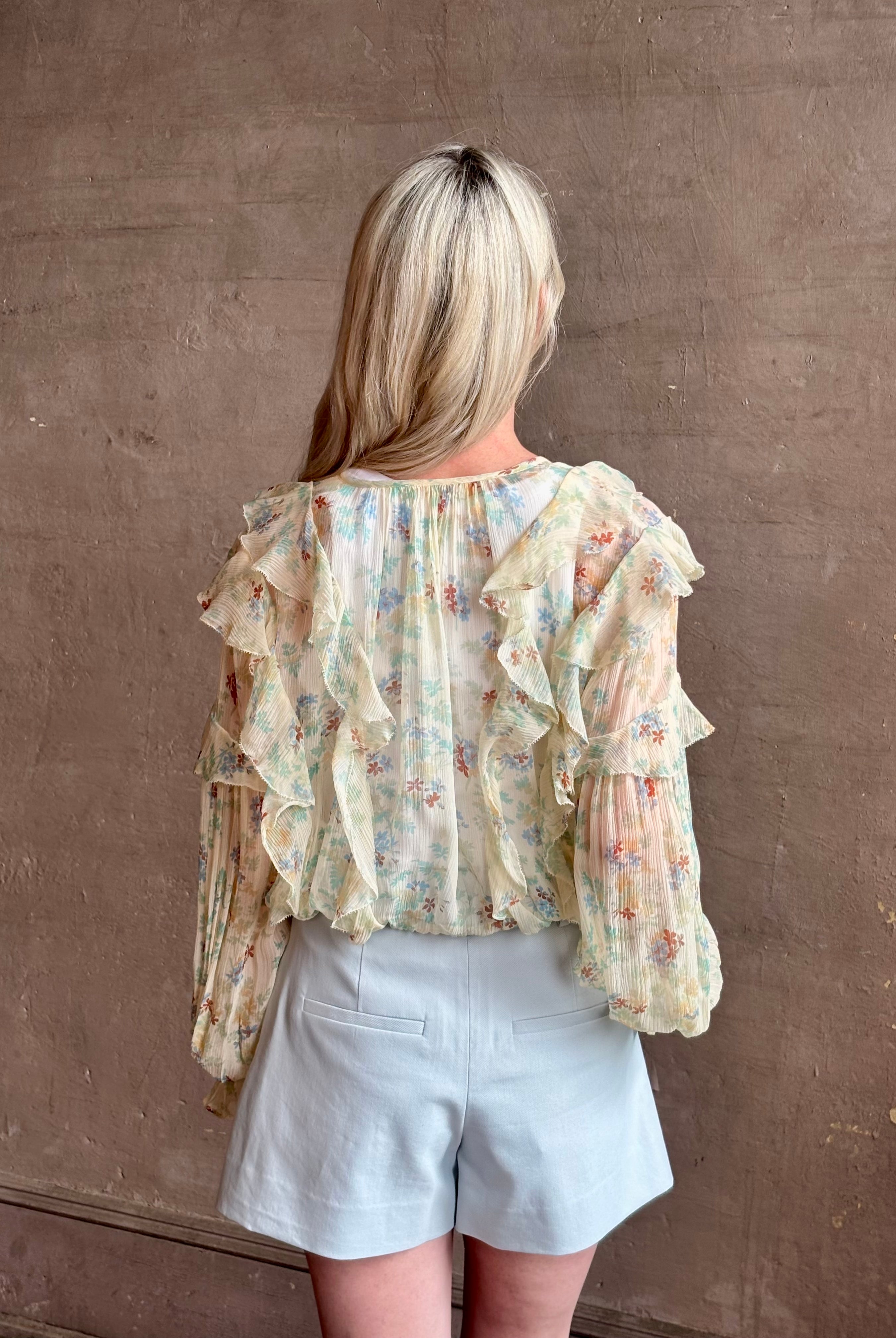Mature woman wearing Delfina Long Sleeve Silk Blouse from Ulla Johnson rendered in a light yellow body adorned with a delicate, scattered tiny floral print with v-neck, drawstring waist, ruffle lined sleeves and linear ruffle details at front and back