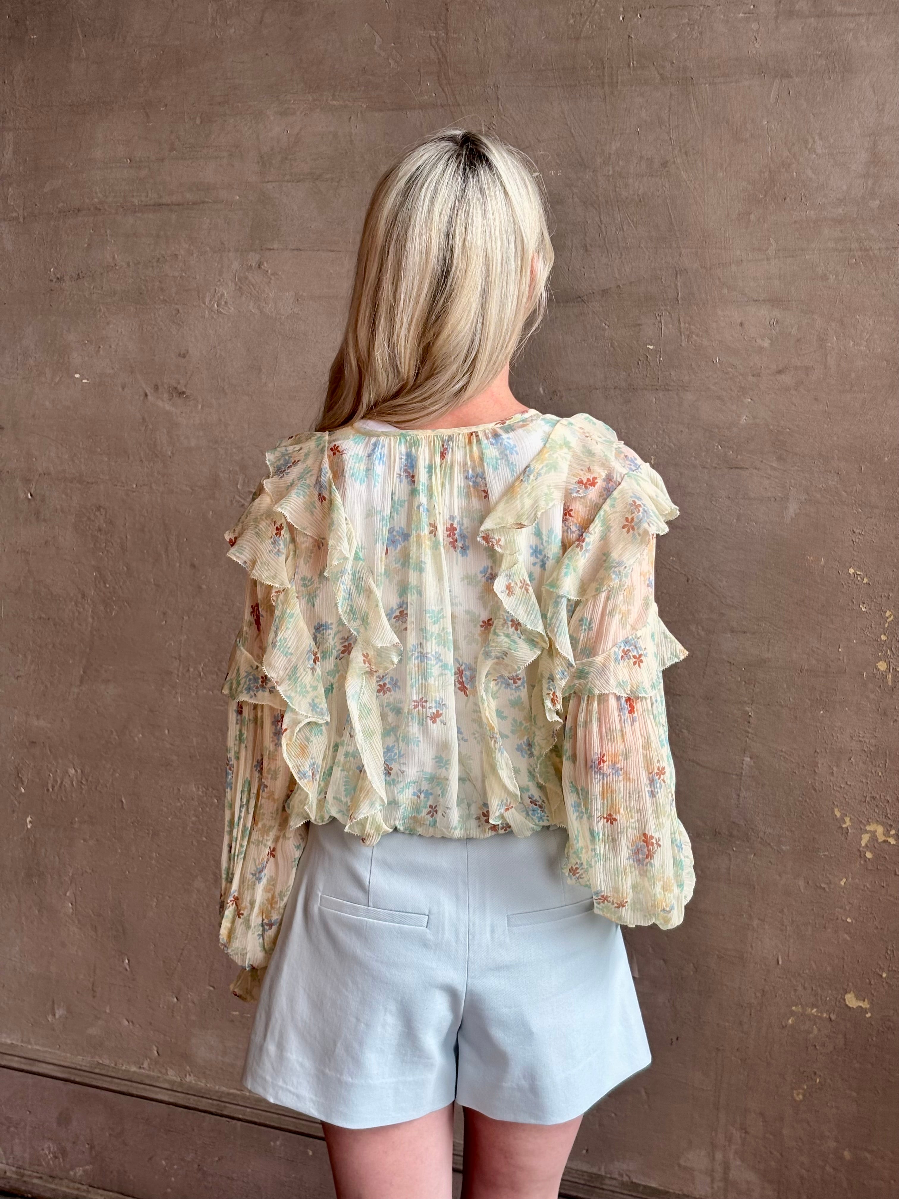 Mature woman wearing Delfina Long Sleeve Silk Blouse from Ulla Johnson rendered in a light yellow body adorned with a delicate, scattered tiny floral print with v-neck, drawstring waist, ruffle lined sleeves and linear ruffle details at front and back