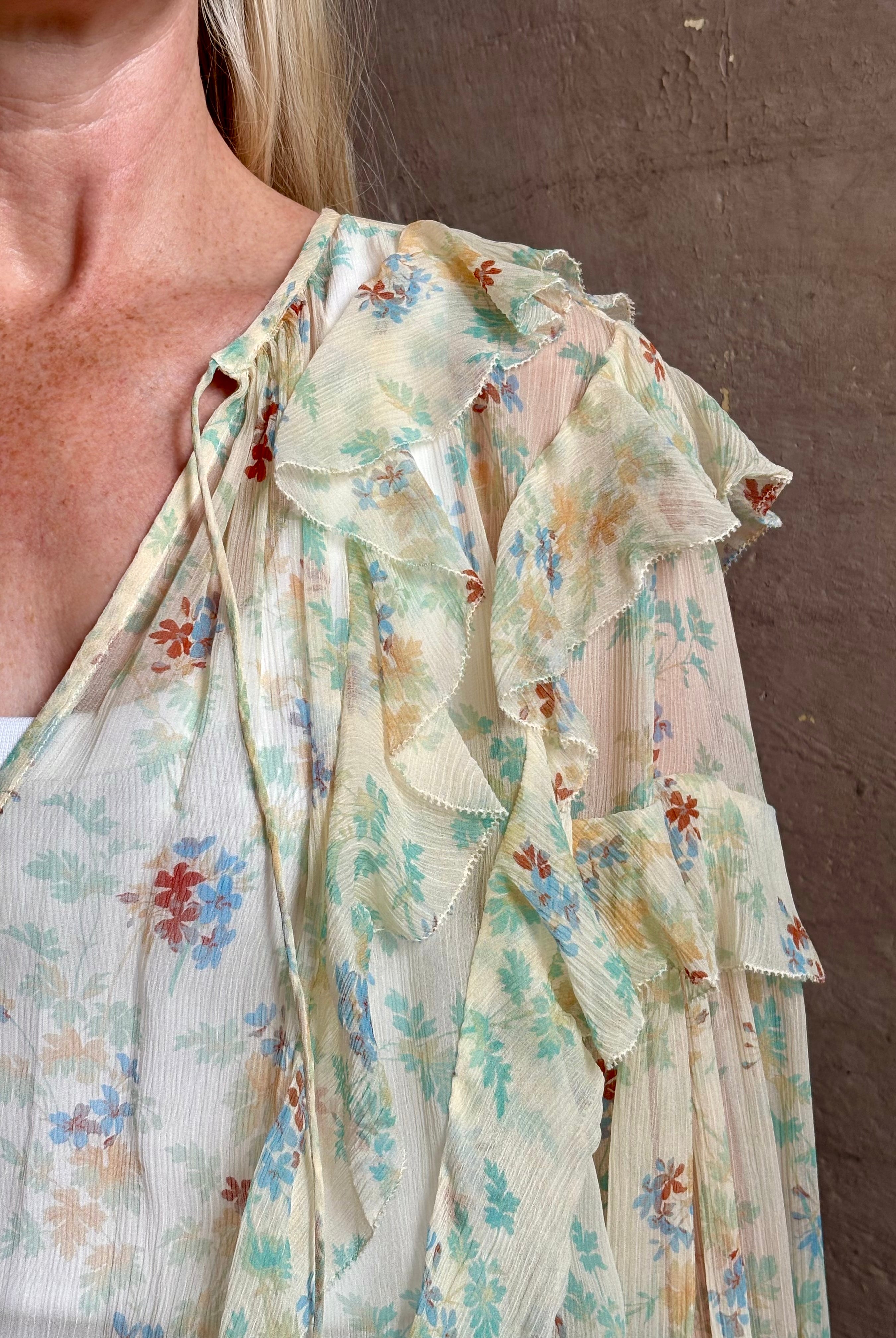 Mature woman wearing Delfina Long Sleeve Silk Blouse from Ulla Johnson rendered in a light yellow body adorned with a delicate, scattered tiny floral print with v-neck, drawstring waist, ruffle lined sleeves and linear ruffle details at front and back