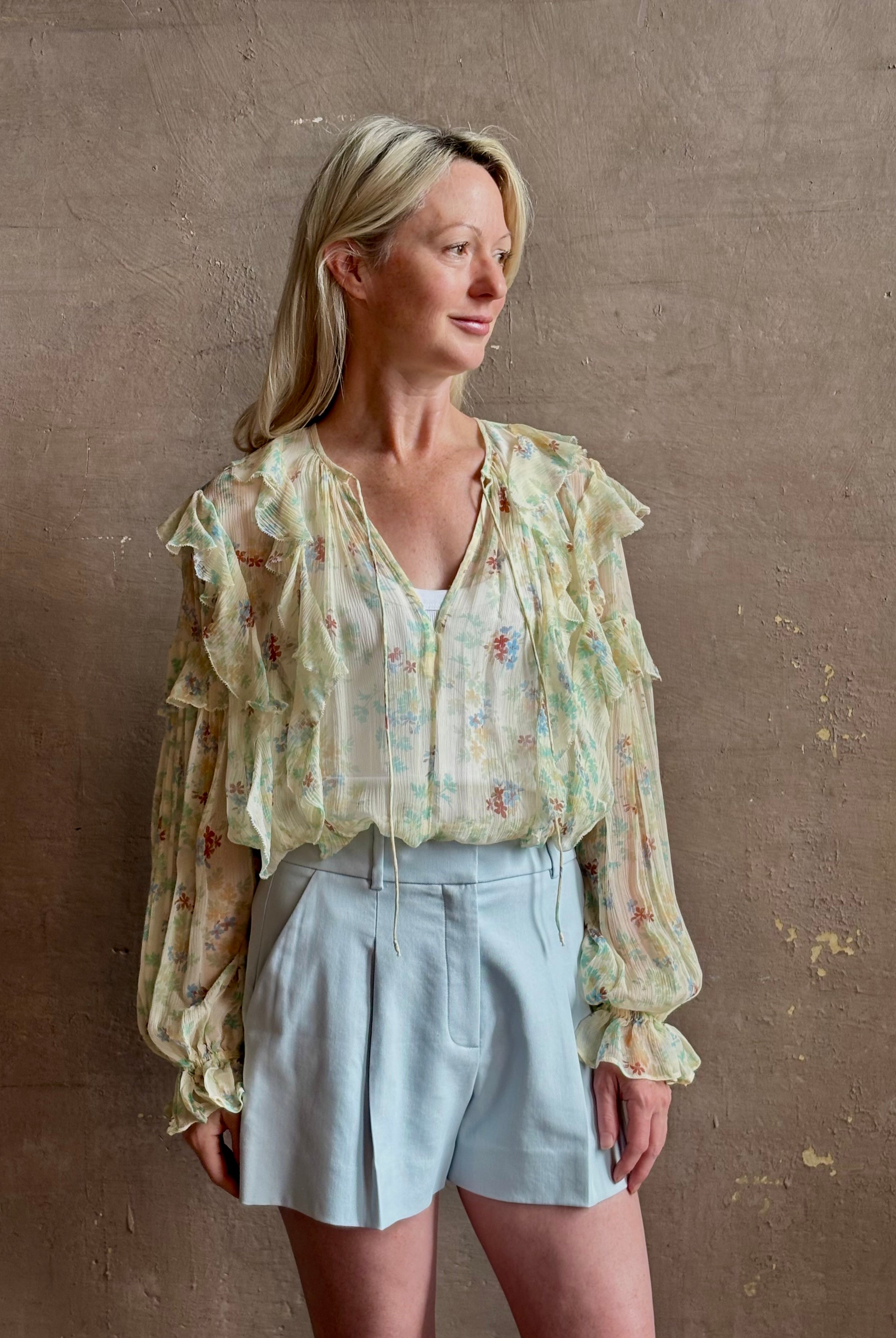Mature woman wearing Delfina Long Sleeve Silk Blouse from Ulla Johnson rendered in a light yellow body adorned with a delicate, scattered tiny floral print with v-neck, drawstring waist, ruffle lined sleeves and linear ruffle details at front and back