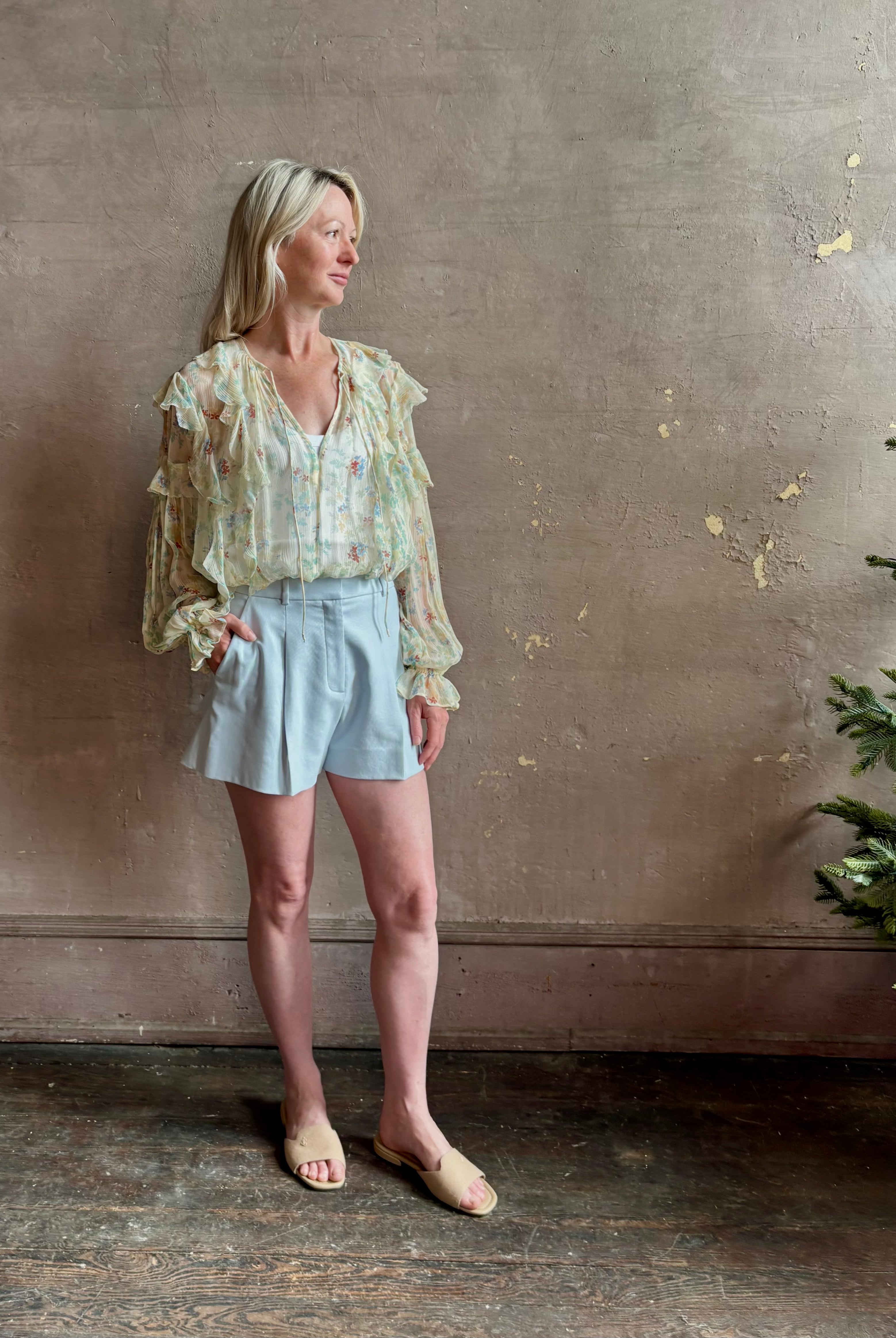 Mature woman wearing Delfina Long Sleeve Silk Blouse from Ulla Johnson rendered in a light yellow body adorned with a delicate, scattered tiny floral print with v-neck, drawstring waist, ruffle lined sleeves and linear ruffle details at front and back