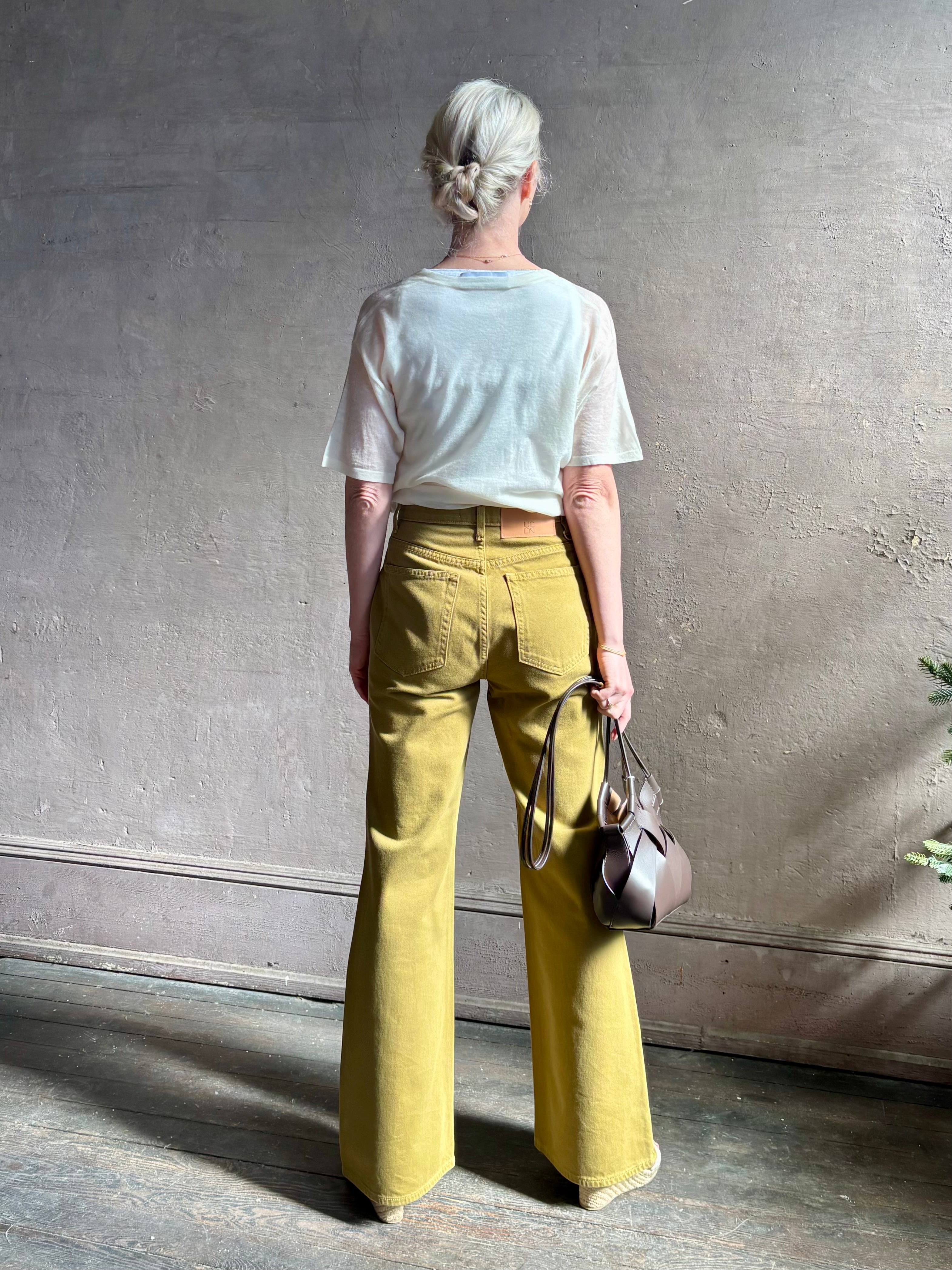 Mature woman wearing luxury Ulla Johnson Lou cotton rigid full length green denim with gold button fly patch pocket