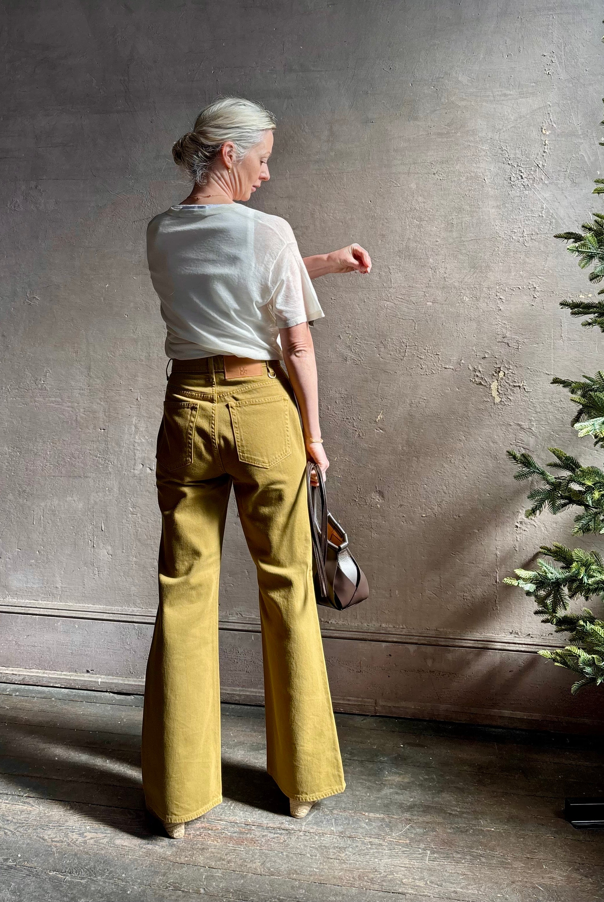 Mature woman wearing luxury Ulla Johnson Lou cotton rigid full length green denim with gold button fly patch pocket