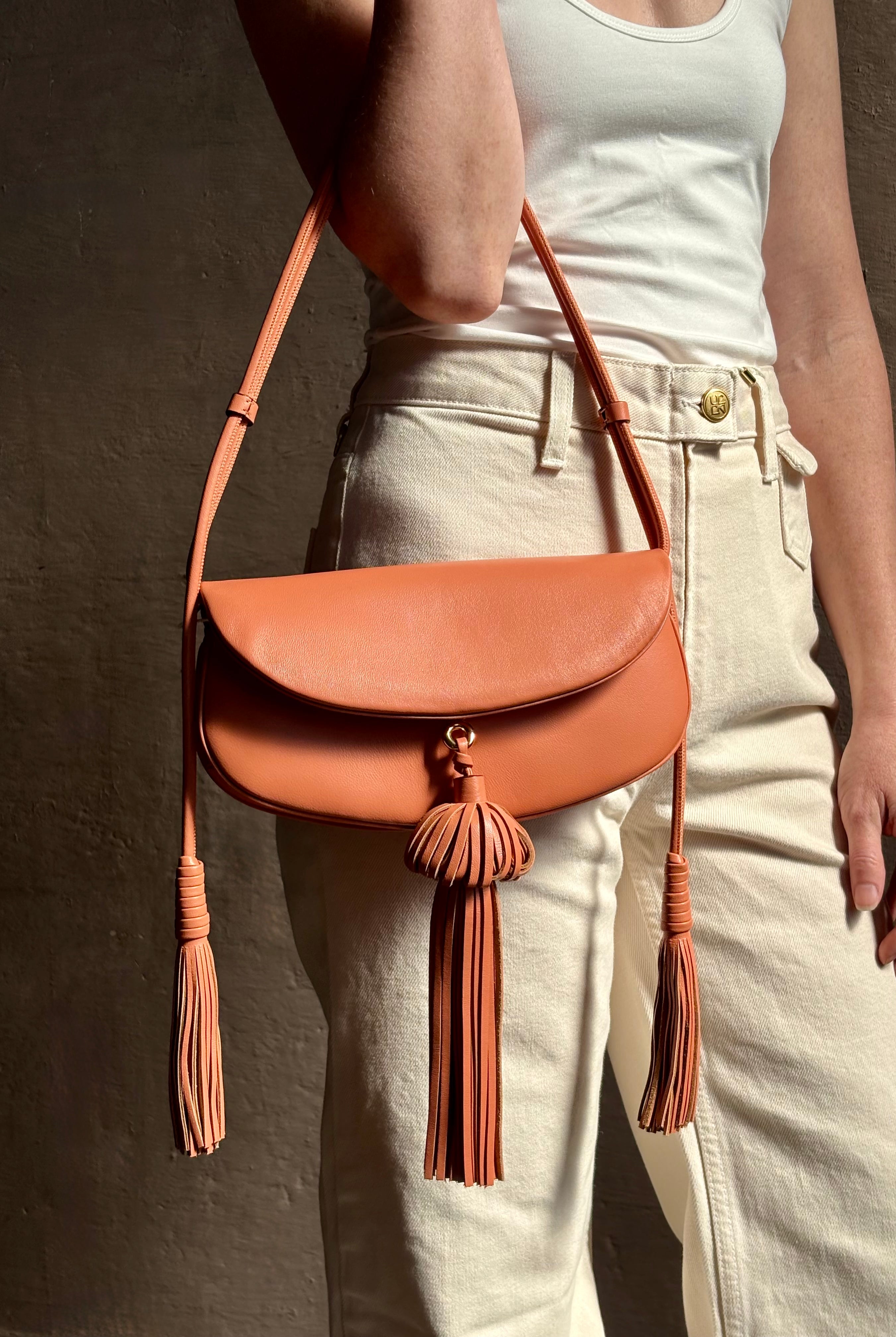 Mature woman with the luxury Italian leather Paloma handbag with Tassel Flap from Ulla Johnson rendered in a warm pink featuring a pull tassel magnetic snap closure and removable adjustable handle that seamlessly converts to a designer clutch