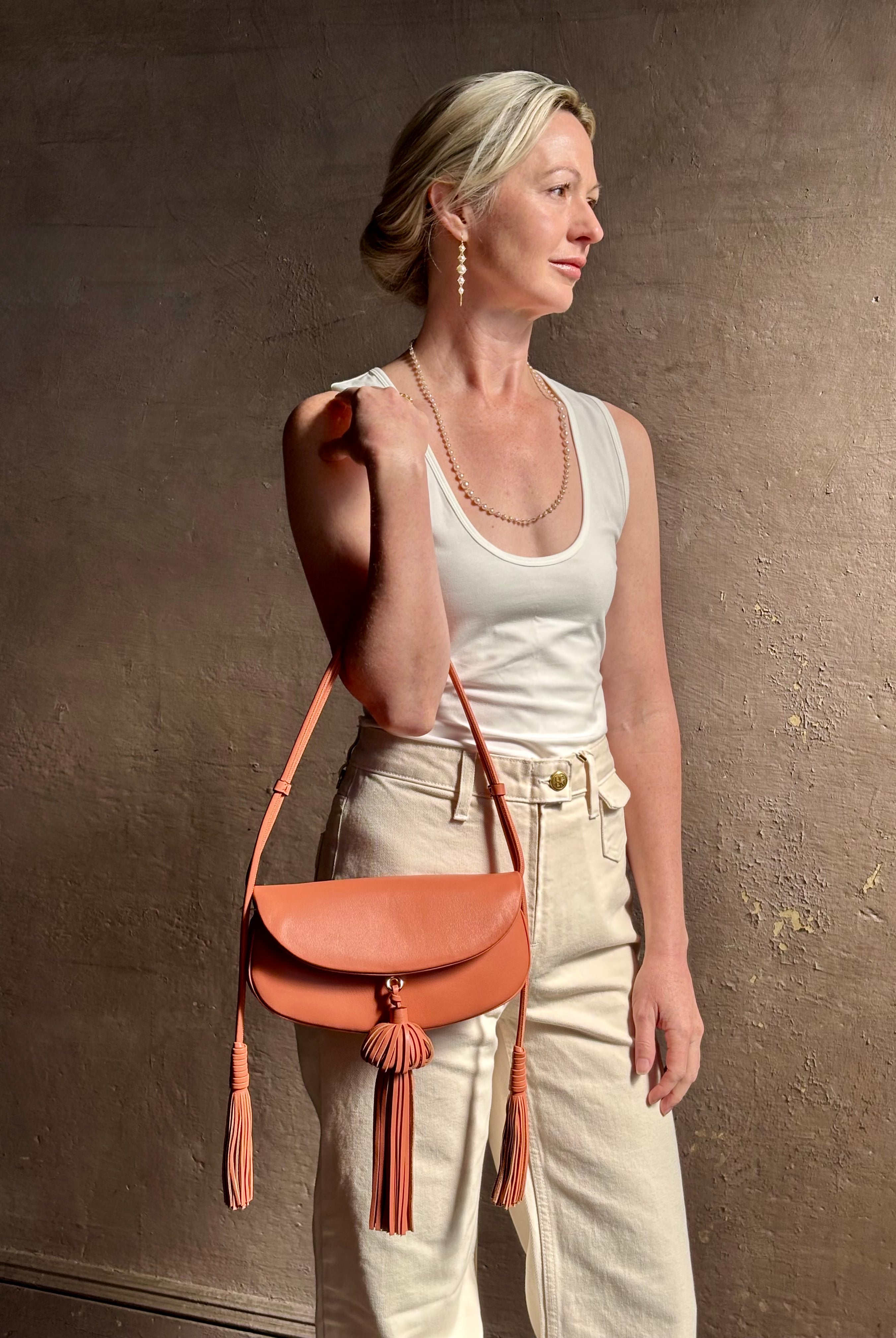 Mature woman with the luxury Italian leather Paloma handbag with Tassel Flap from Ulla Johnson rendered in a warm pink featuring a pull tassel magnetic snap closure and removable adjustable handle that seamlessly converts to a designer clutch