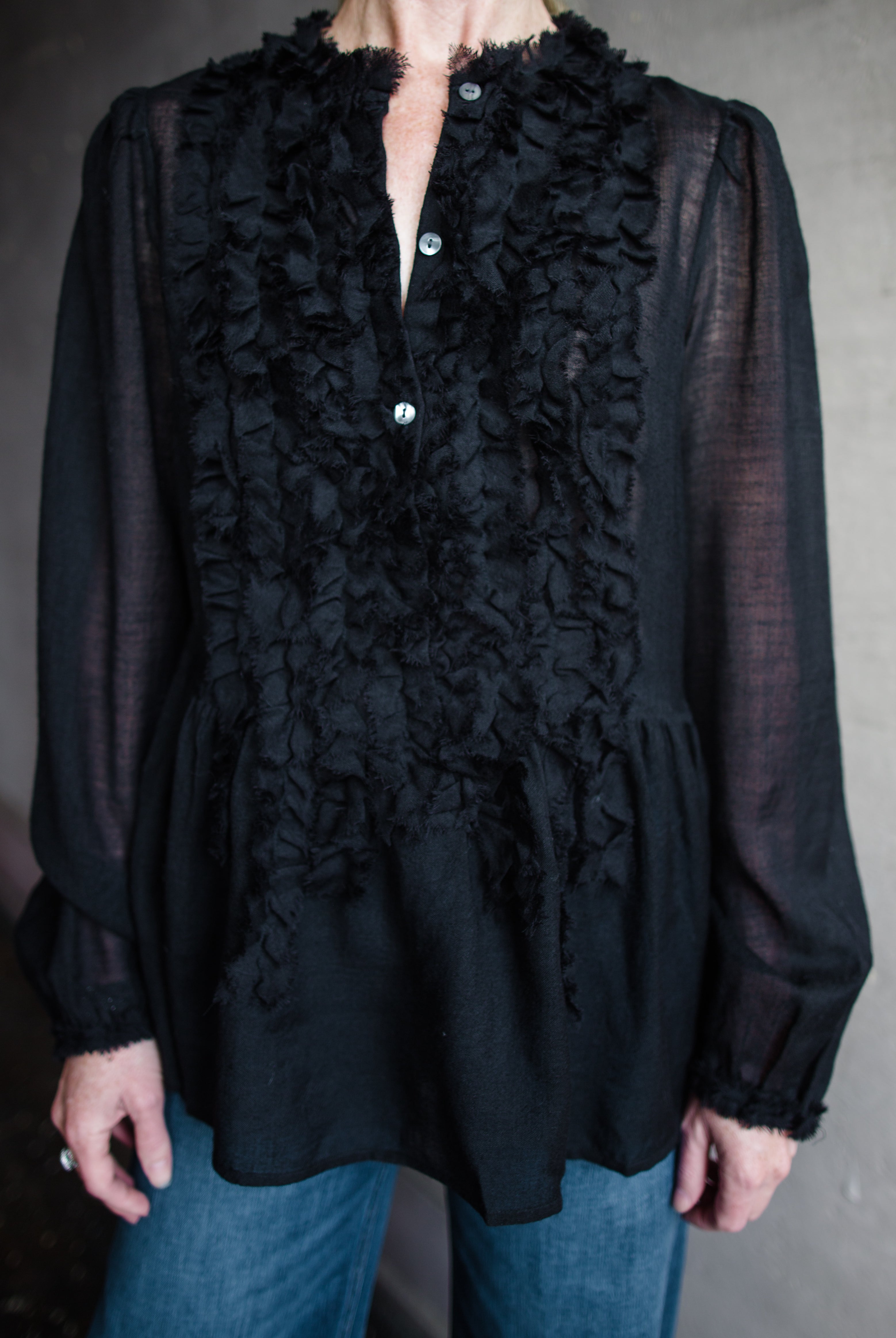 Image features woman wearing The Cosima Tunic by Ulla Johnson in Noir features a button down v neckline with rows of ruffles and iridescent sea shell buttons.